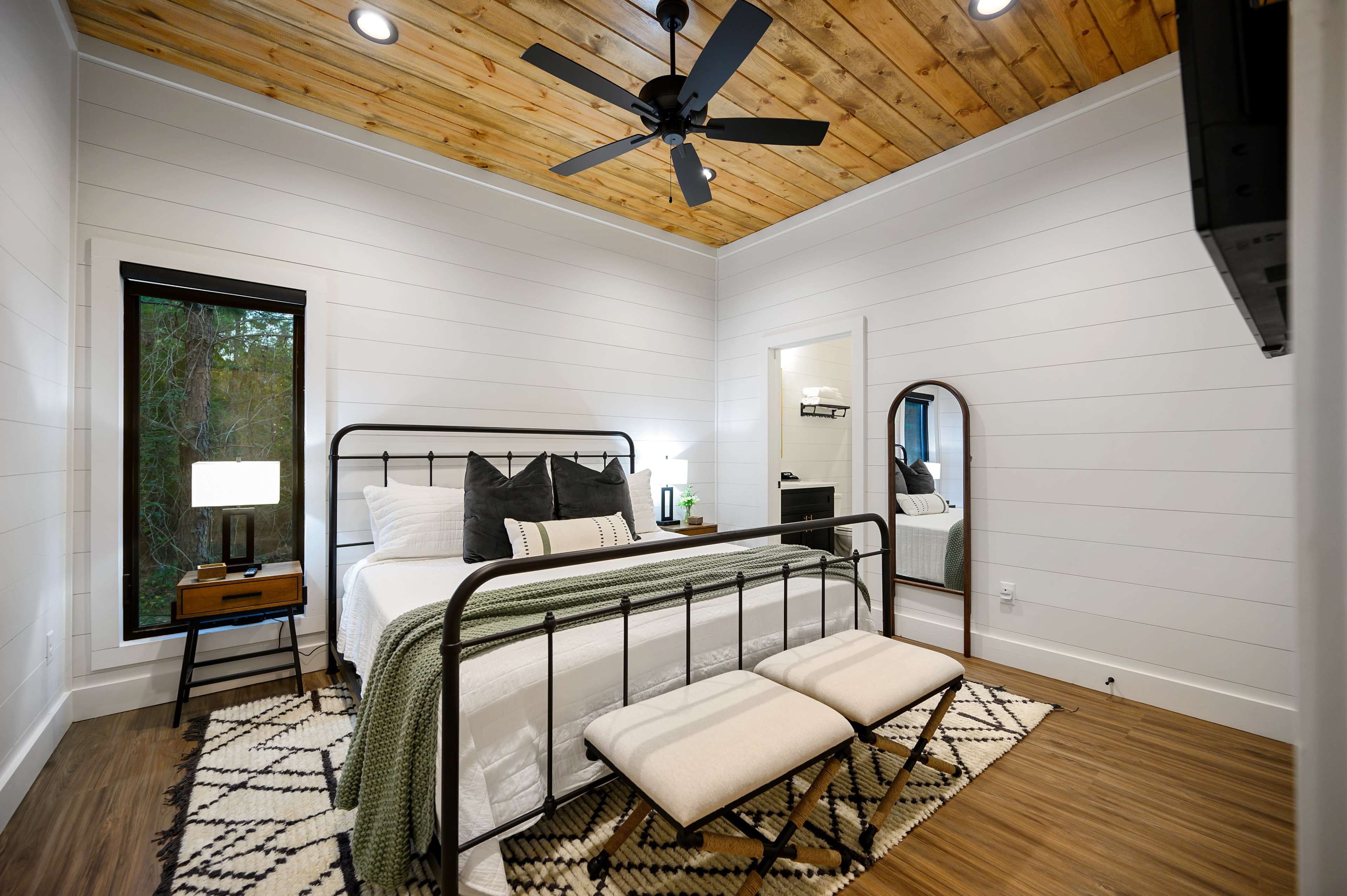 A modern bedroom featuring a metal bed frame, a wooden ceiling, and a cozy area rug with a mirror and window in the background.