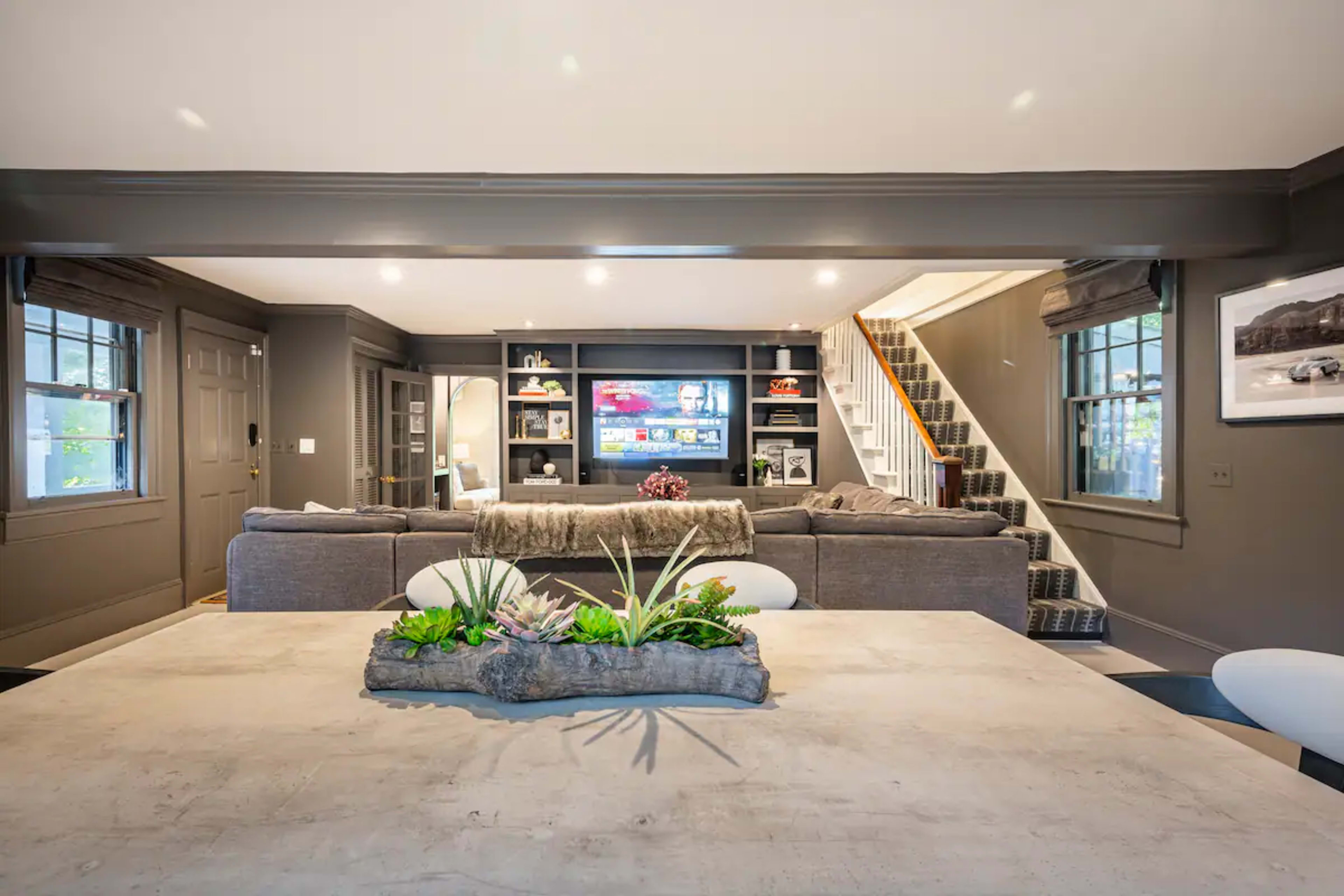 The image shows a modern living room with a large gray sofa, a television mounted on the wall, and a dining table with a plant centerpiece.