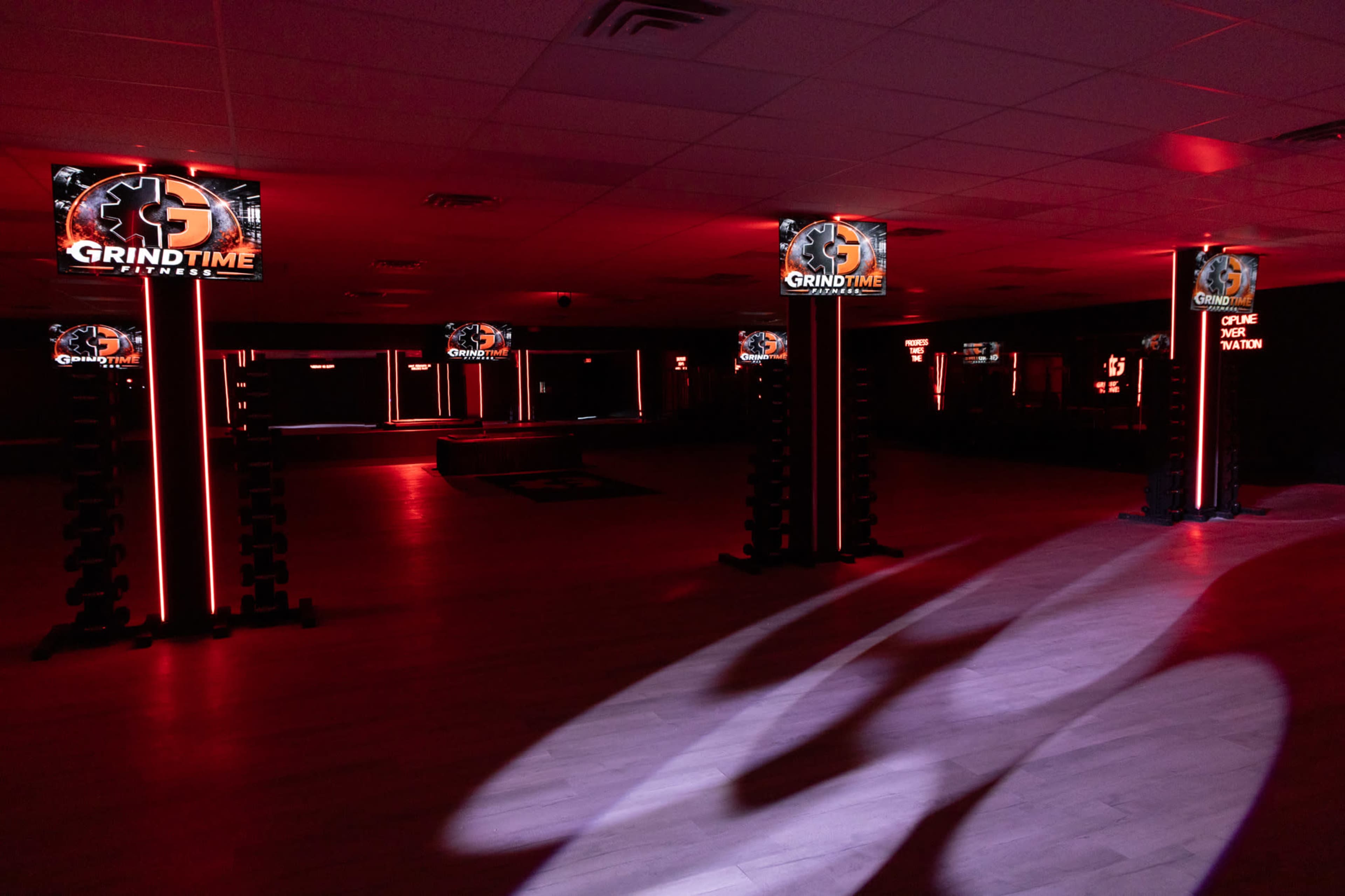 The image shows a dimly lit fitness studio featuring several illuminated displays promoting "GRINDTIME" and equipped with weights on the floor.