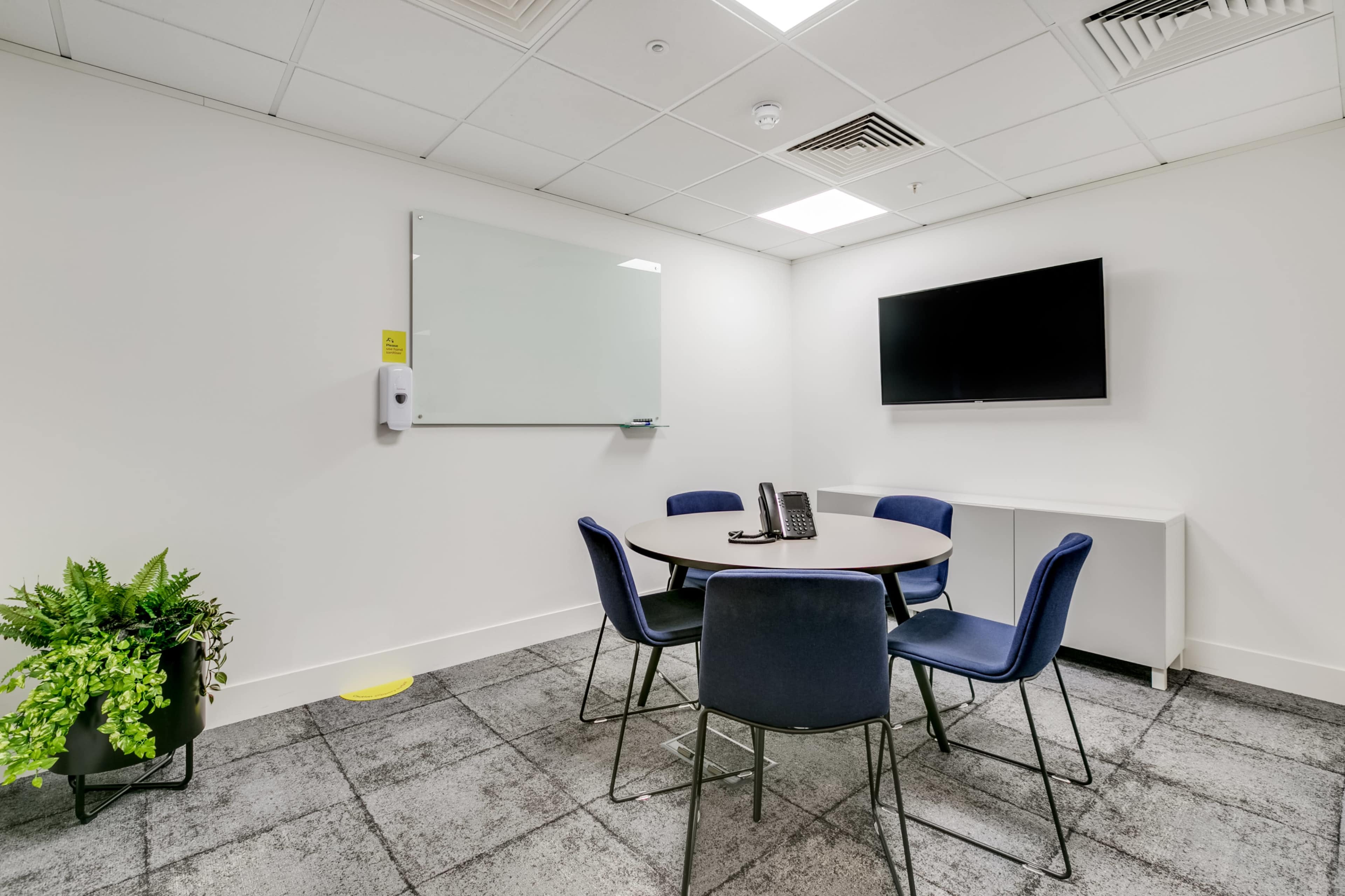 A small meeting room features a round table with four chairs, a wall-mounted television, a whiteboard, and a plant in a corner.