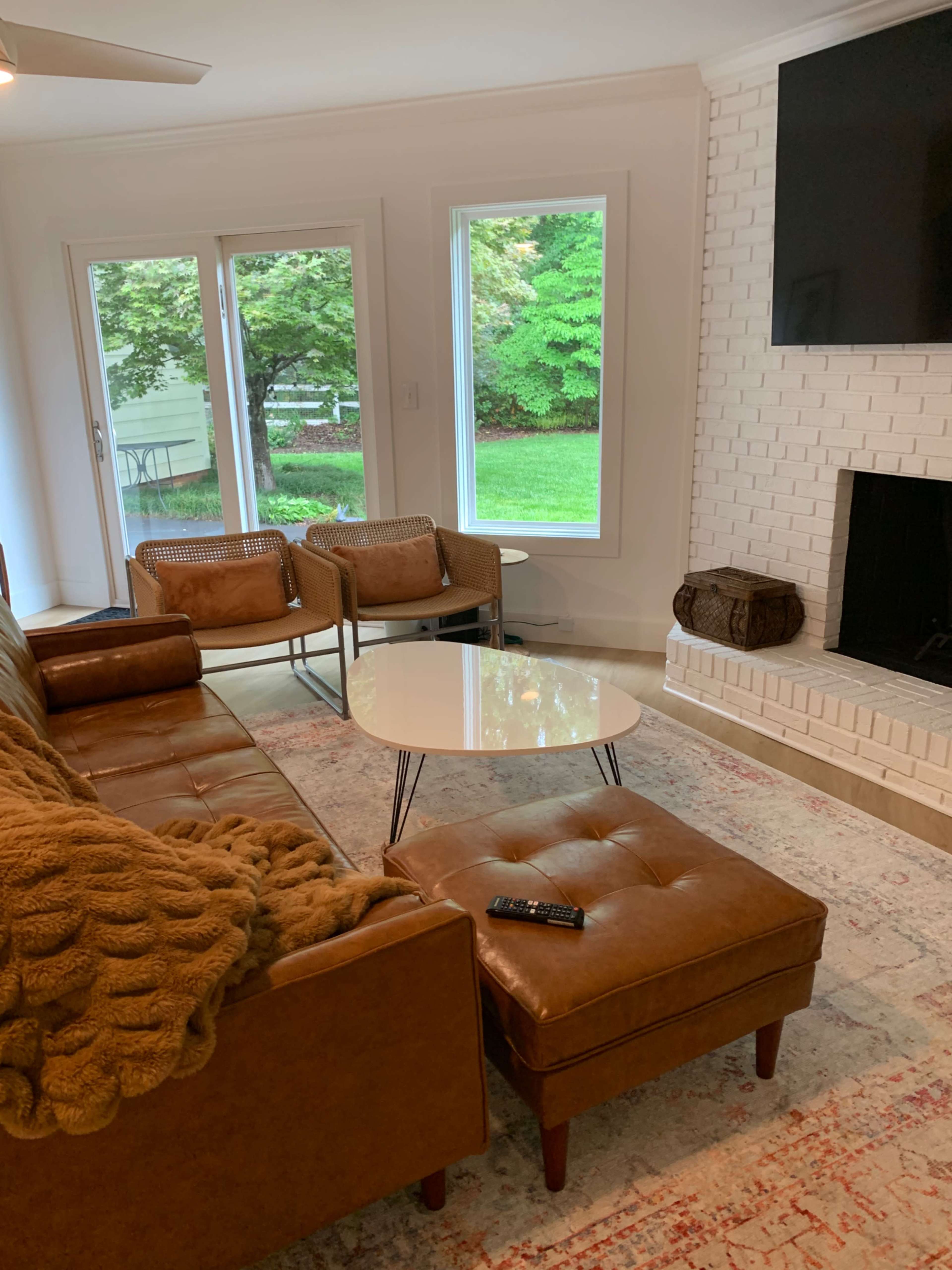 A modern living room features a brown leather sectional sofa, two woven chairs, and a round coffee table in front of a fireplace and large windows overlooking a green yard.