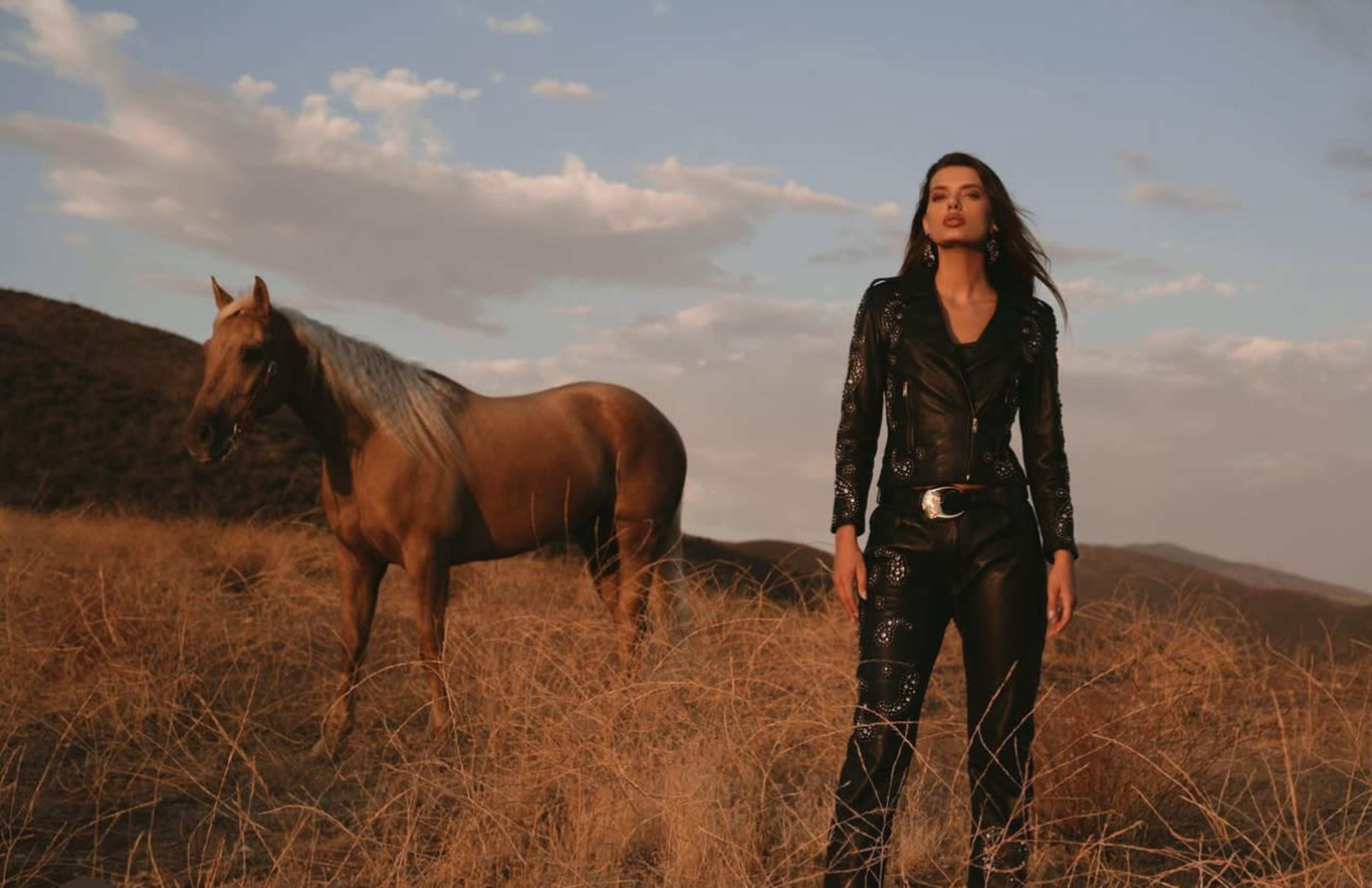 A woman in a leather outfit stands in a grassy field beside a horse under a cloudy sky.