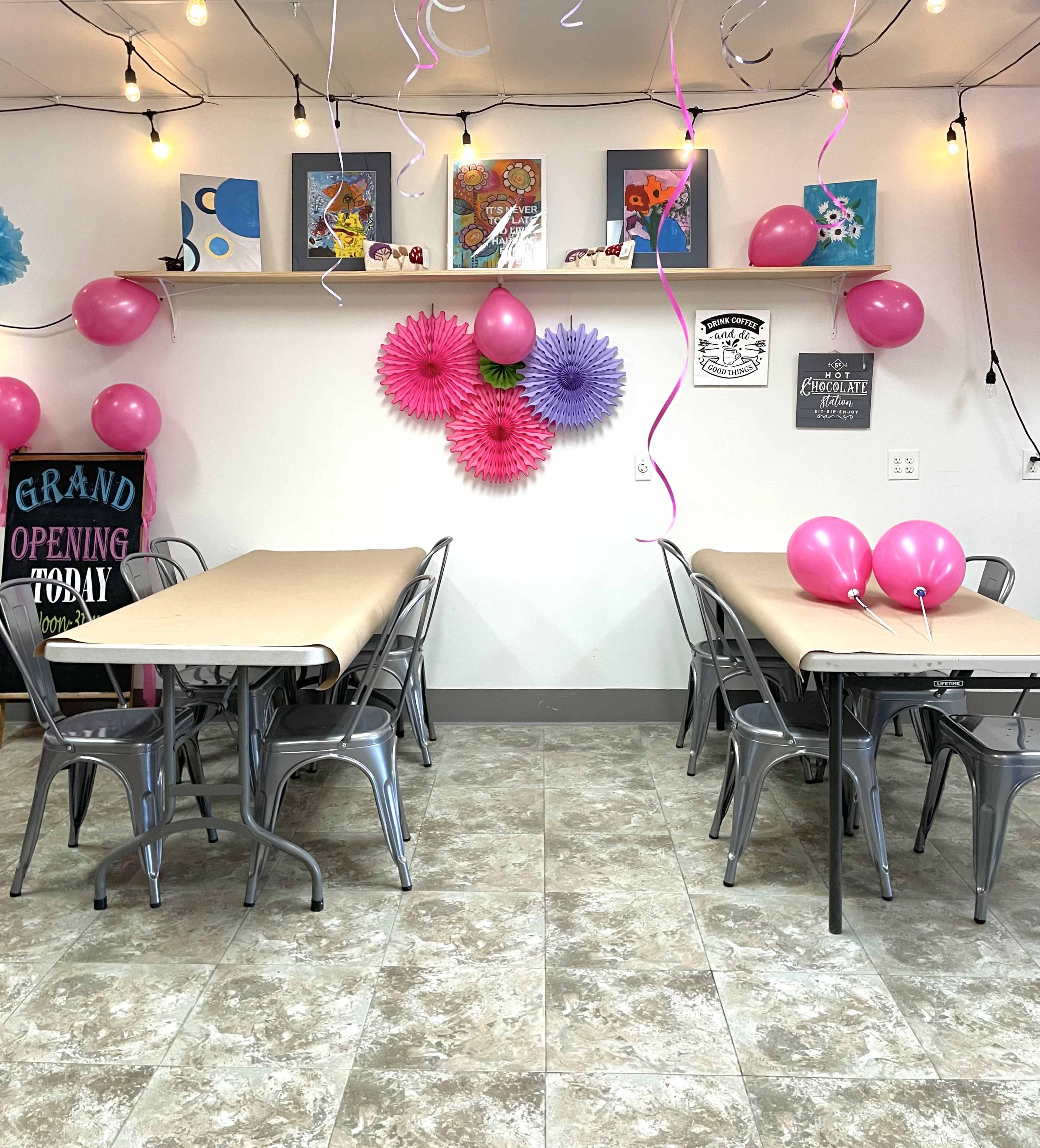 The image shows a decorated event space with two tables, pink balloons, and colorful paper flowers on the wall, set for a grand opening celebration.