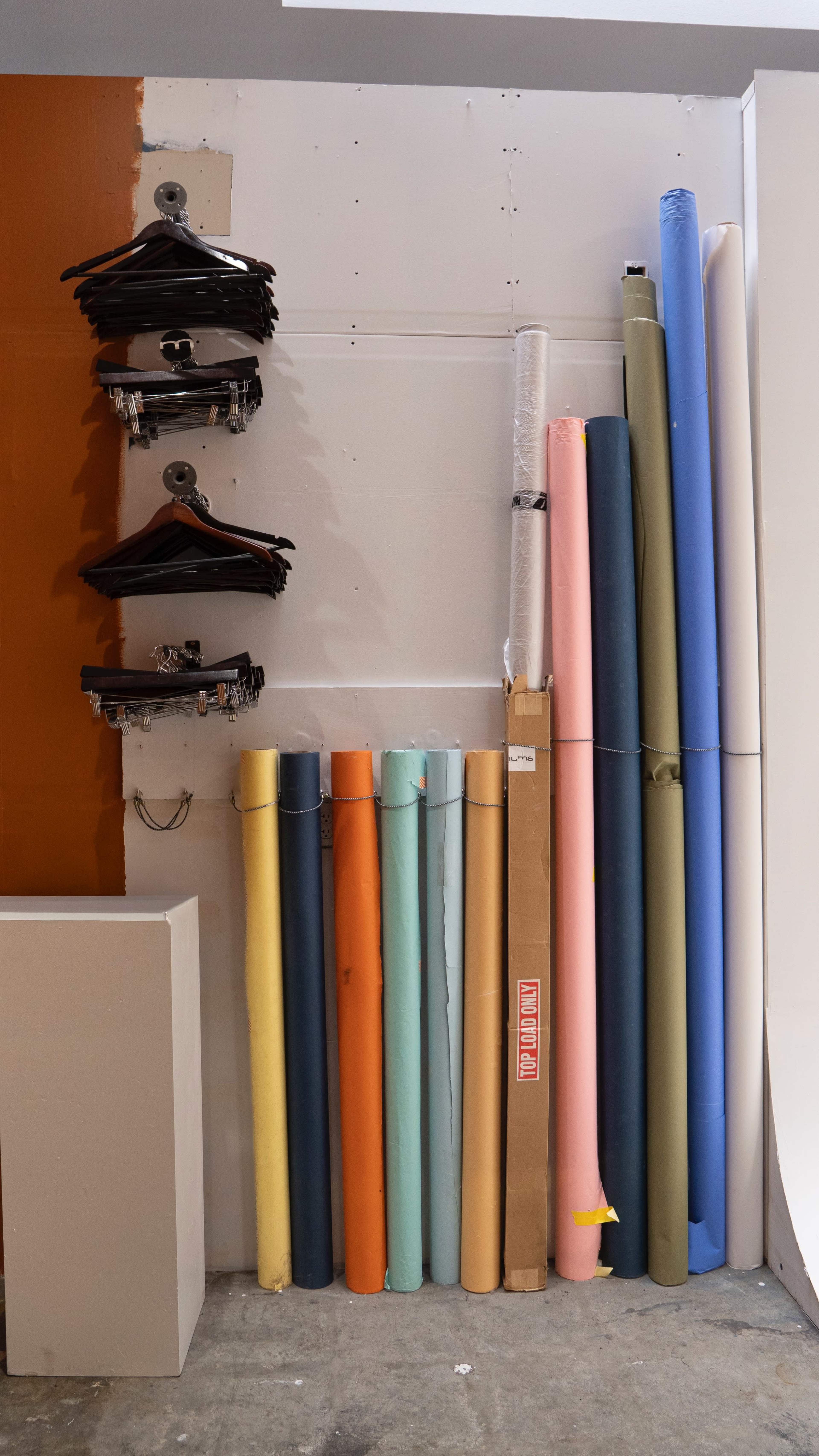 The image shows a wall with various colored rolls of paper stacked vertically beside a set of hangers on hooks.