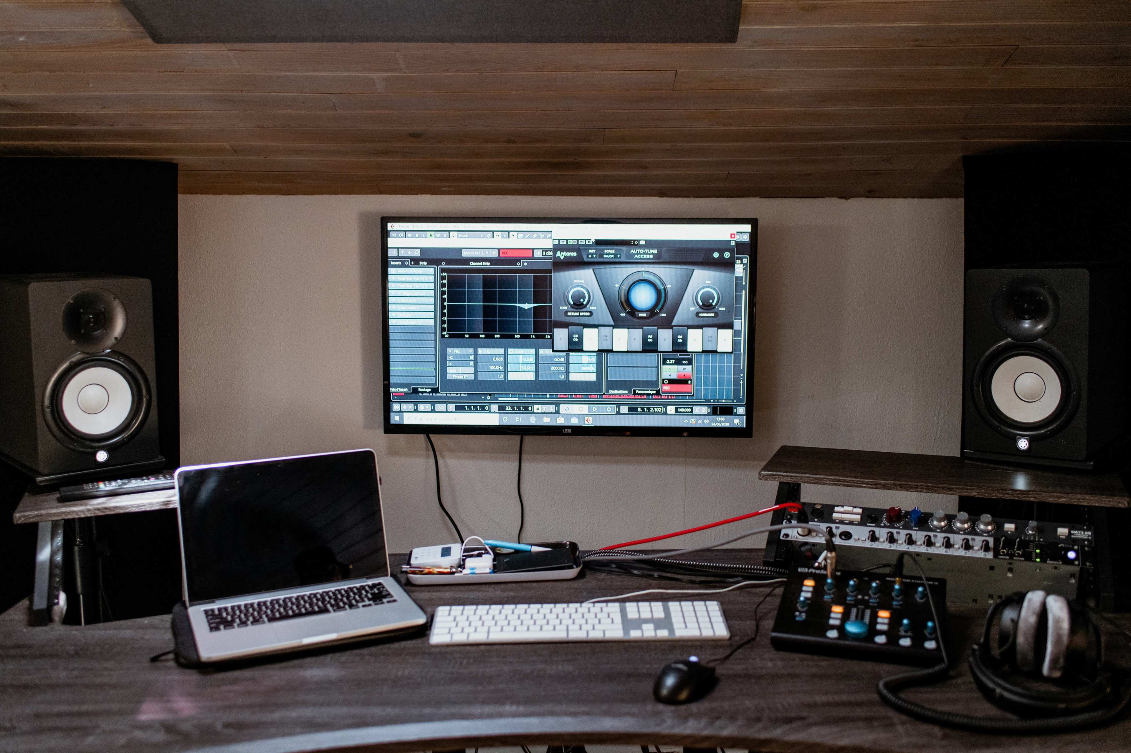 A music production workspace features a large monitor displaying software, flanked by two studio monitors, with a laptop and audio equipment on the desk.