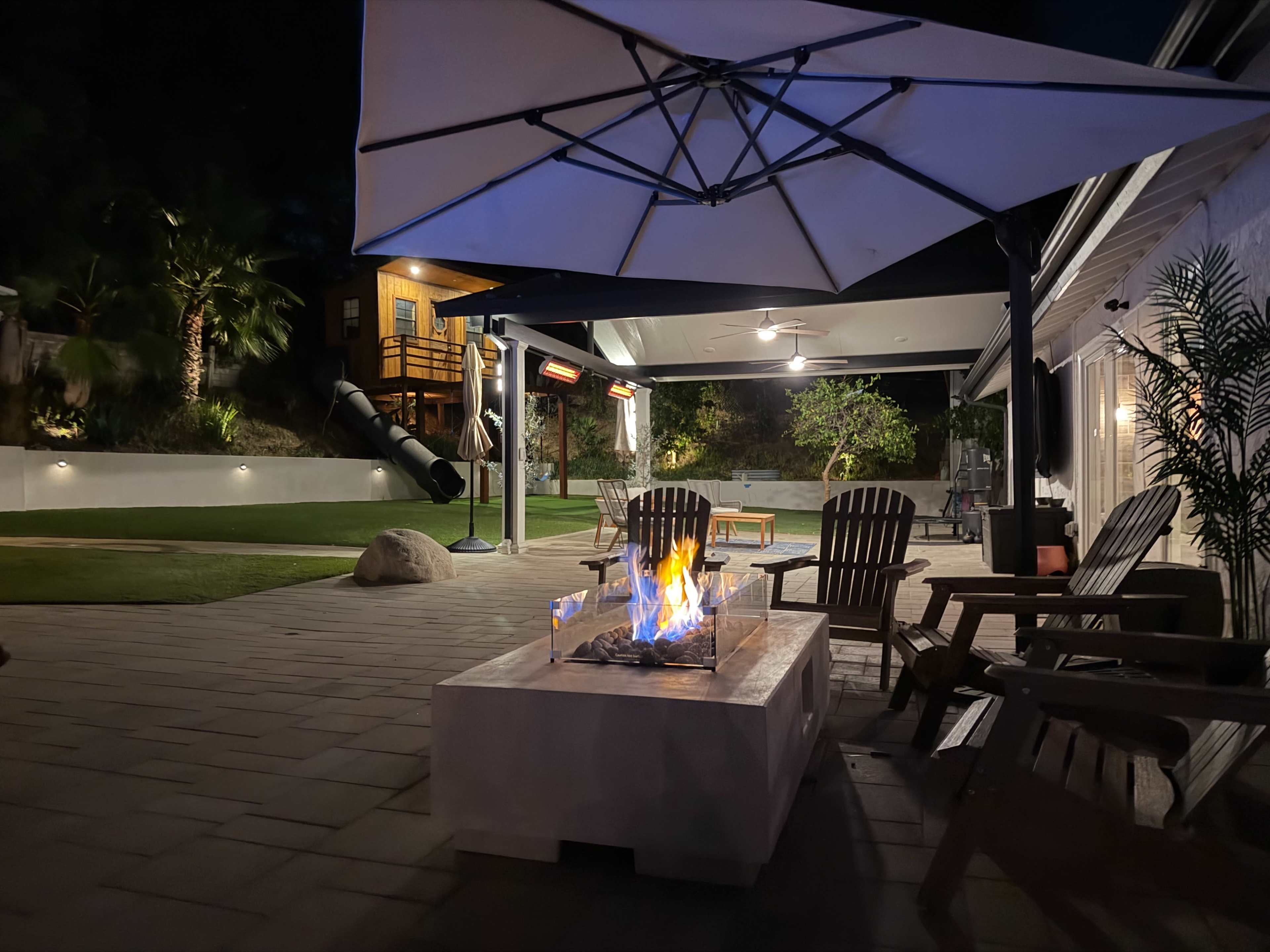 A backyard with a fire pit surrounded by Adirondack chairs, a large umbrella overhead, and a slide in the background near a playhouse, all illuminated at night.