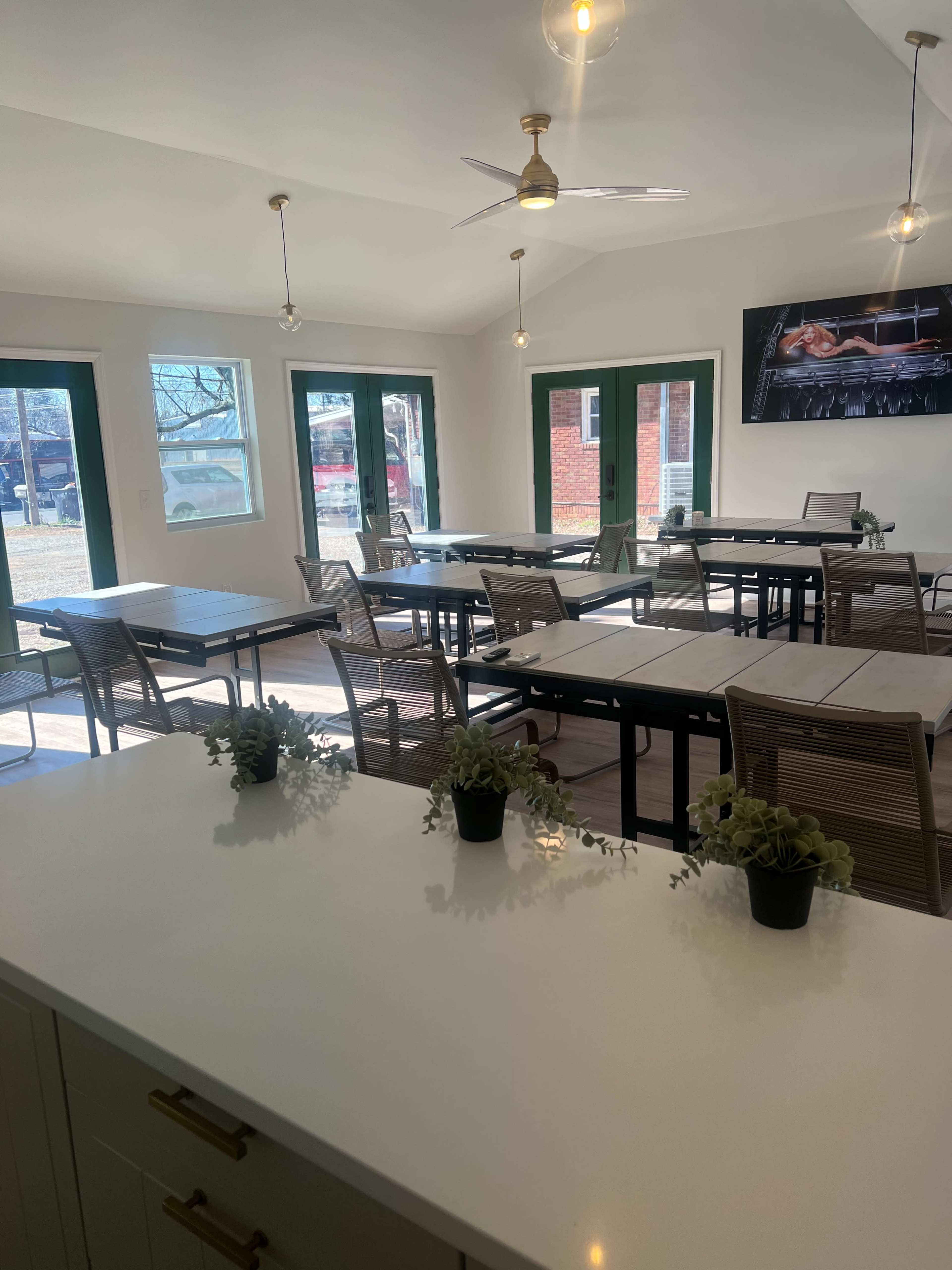 The image shows a modern classroom or meeting space with several tables and chairs arranged neatly, large windows allowing natural light, and potted plants on the tables.