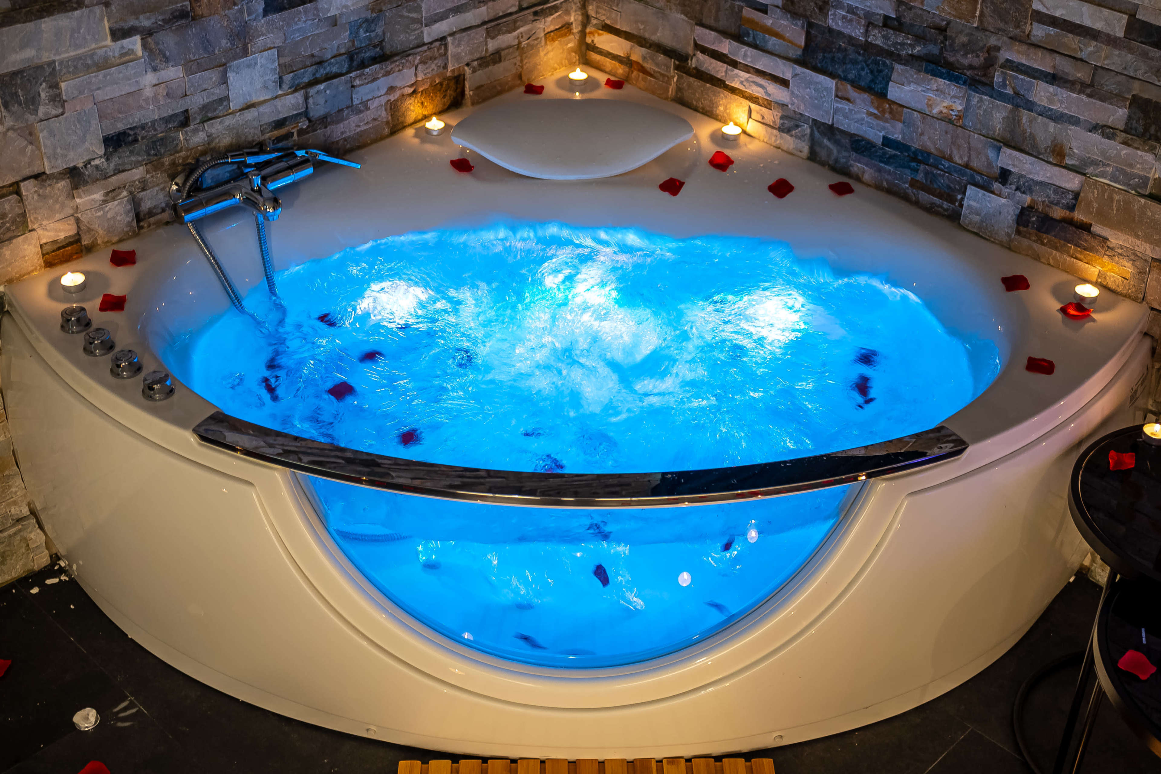 A corner jacuzzi tub filled with blue-lit water, surrounded by rose petals and candles against a stone wall.