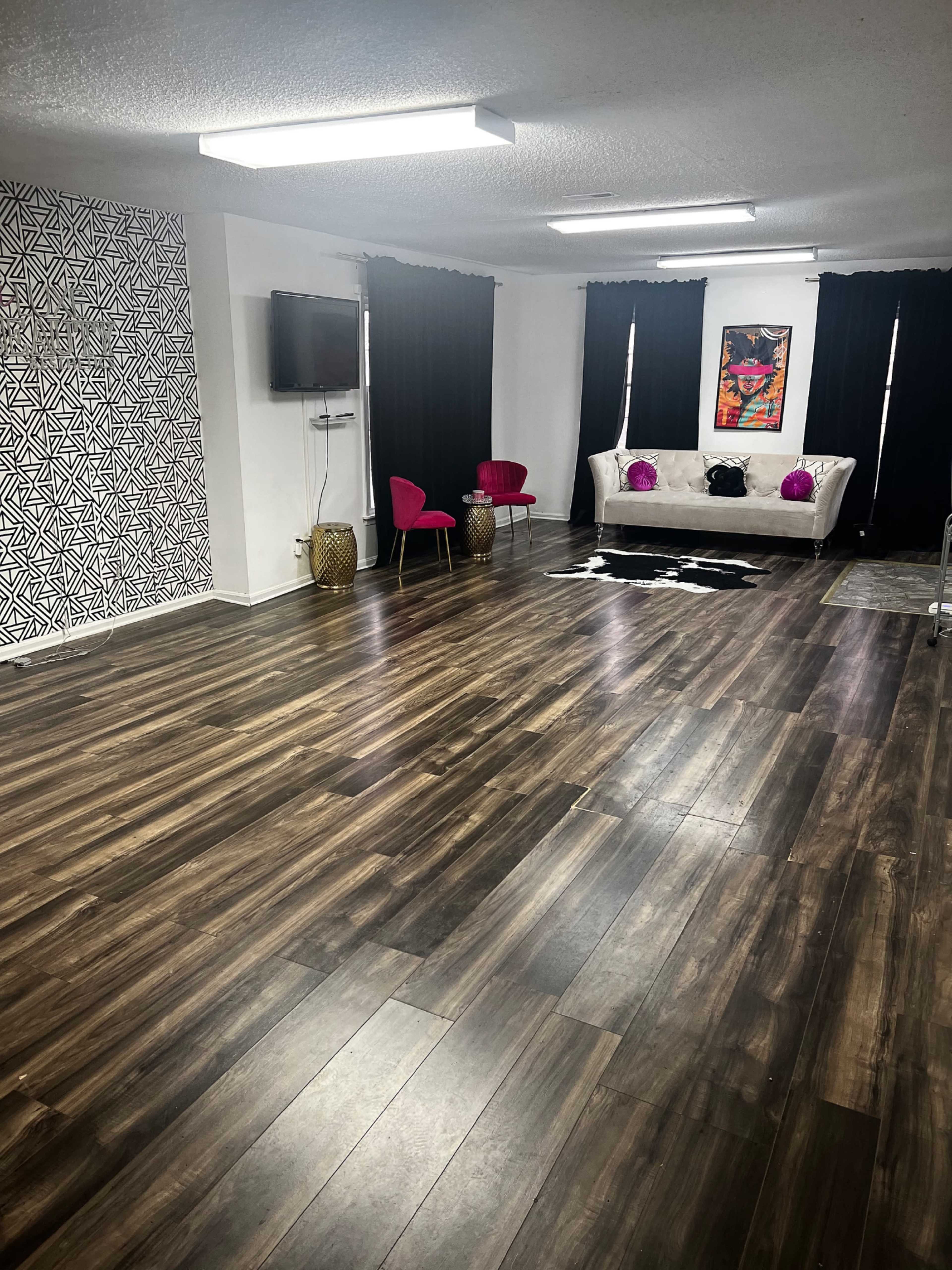 A spacious room with dark wooden flooring, a patterned accent wall, a television mounted on one side, and a white couch with colorful chairs.