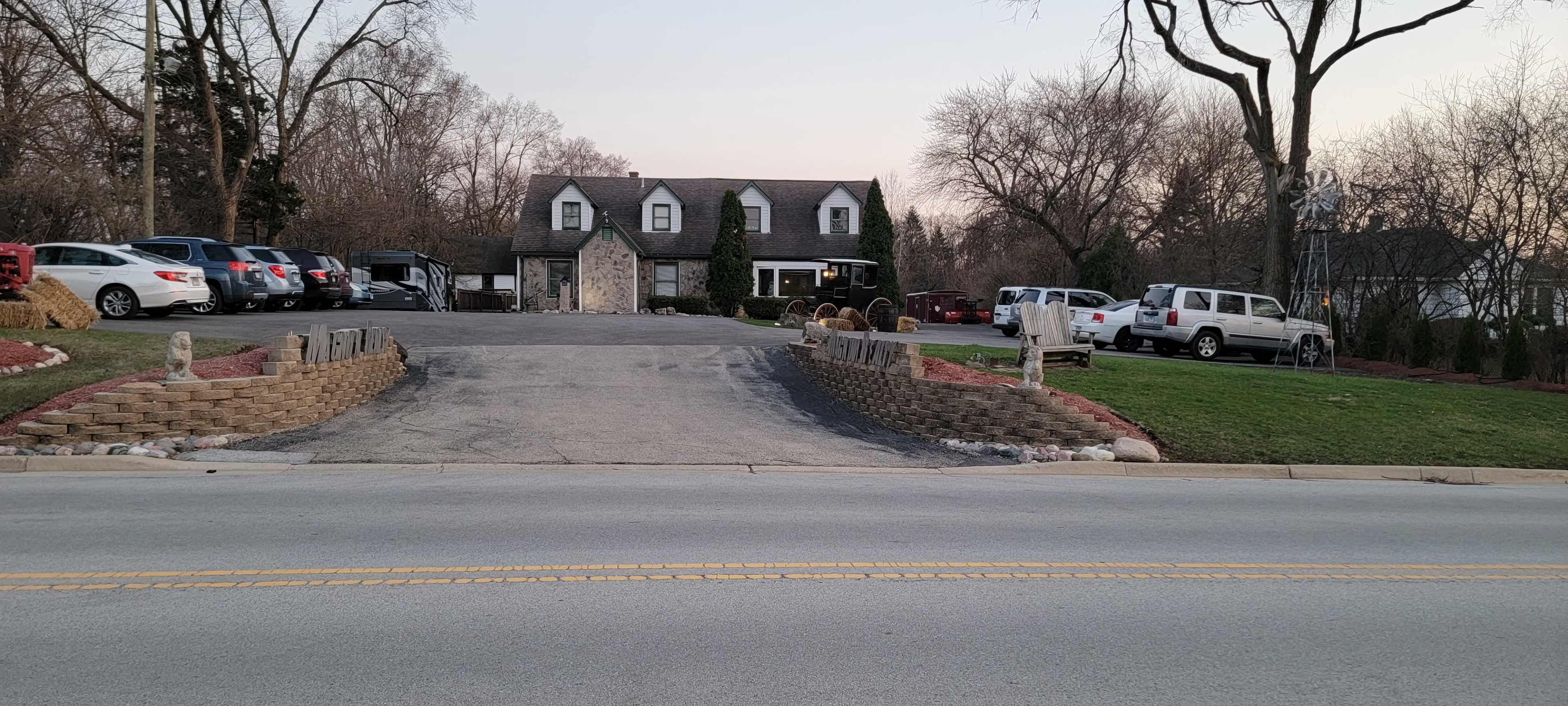 A two-story house with a dark roof, surrounded by several parked vehicles, situated at the end of a winding driveway, with grassy areas and trees in the background.