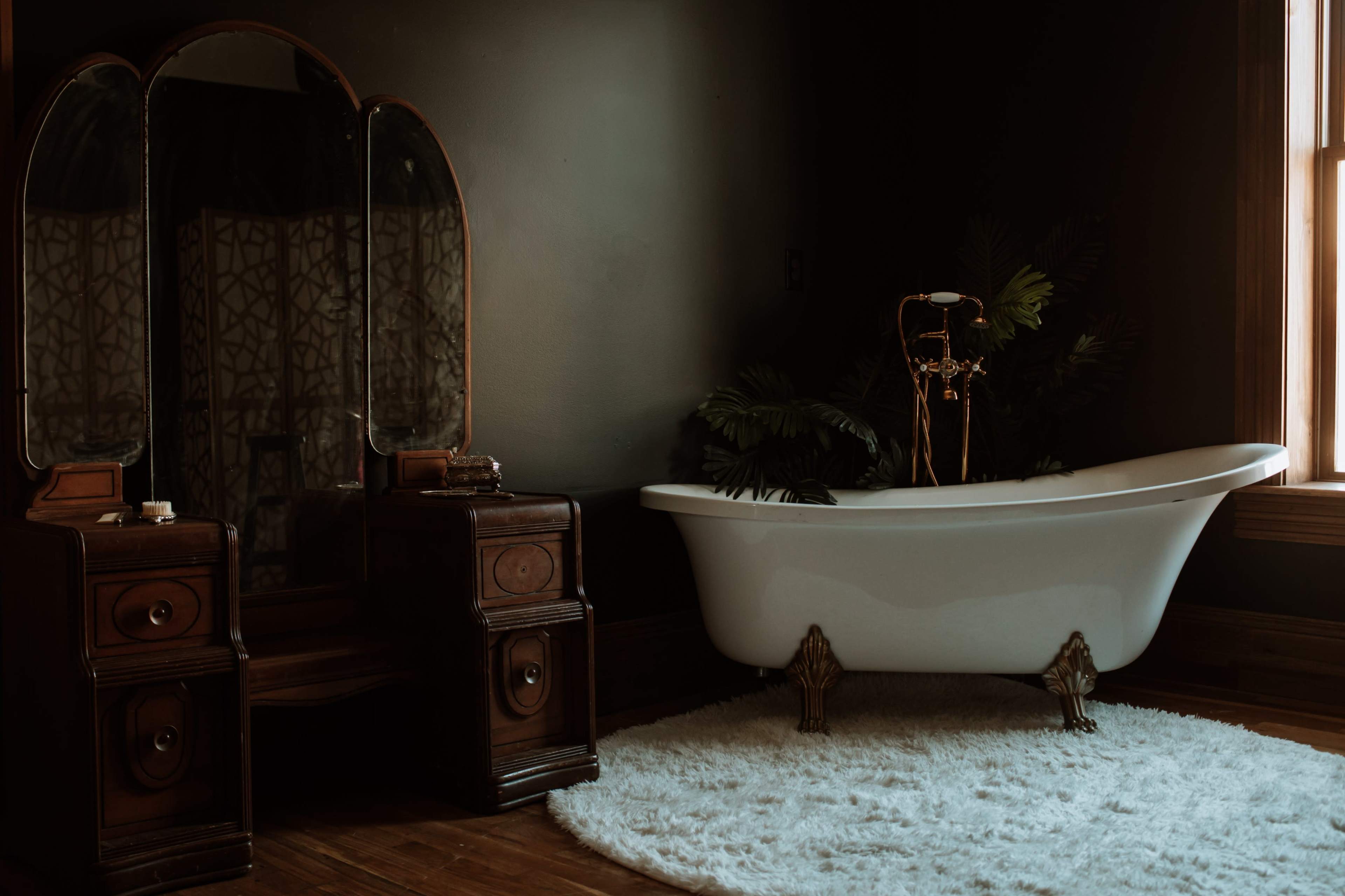 A vintage bathroom features a freestanding white bathtub beside a wooden vanity with a large mirror, all set against a dark wall.