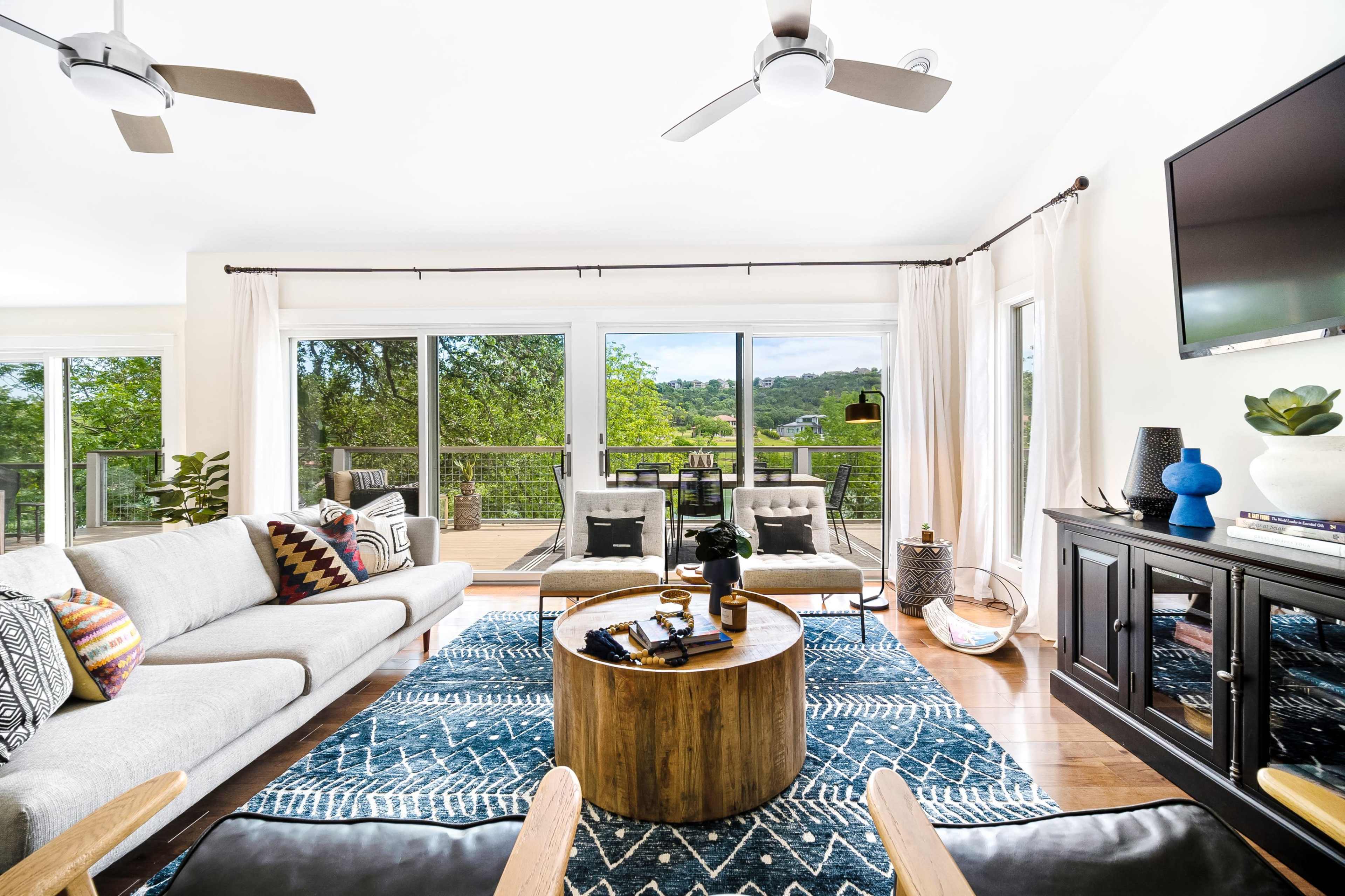 The image shows a bright living room with a gray sofa, a circular wooden coffee table, and large glass doors opening to a deck with a view of greenery.