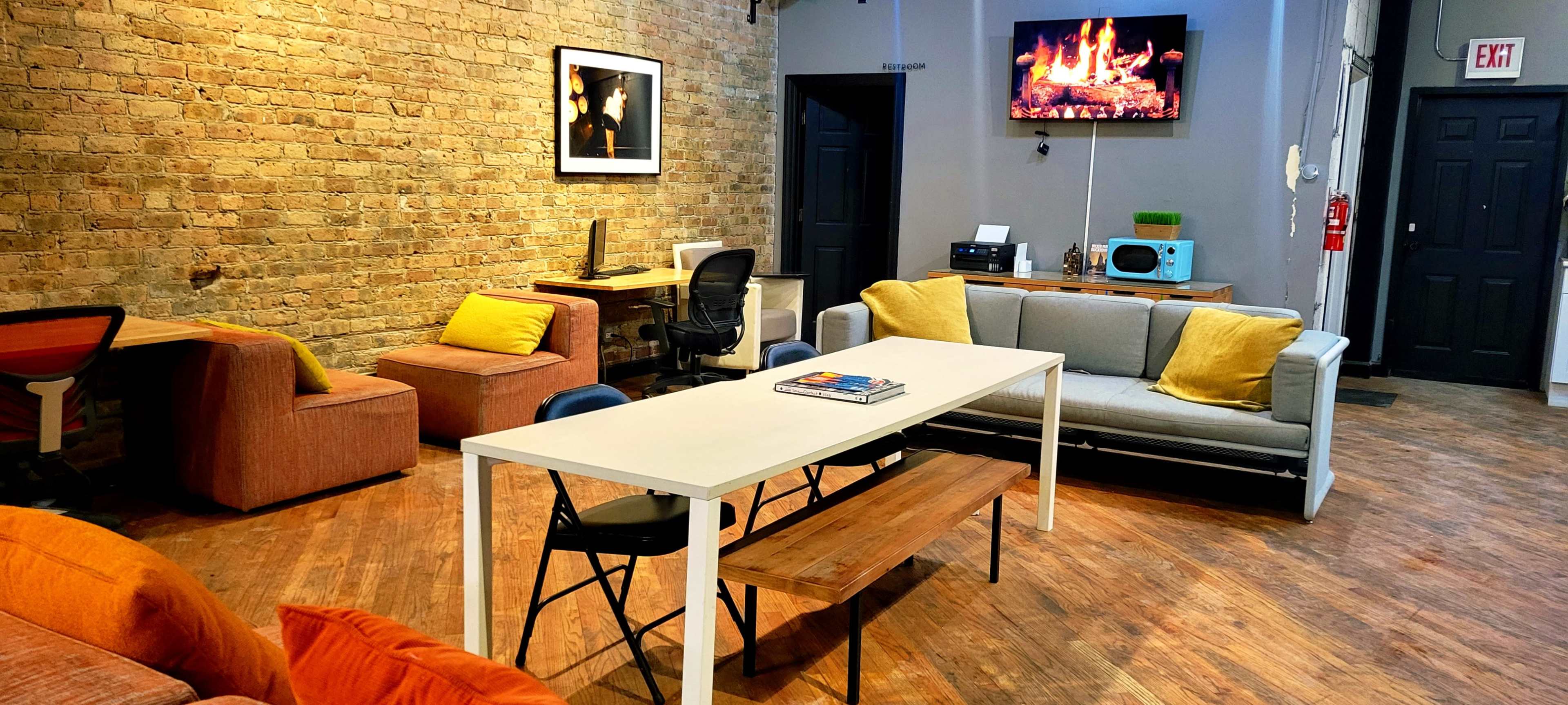 The image shows a spacious, modern lounge area with a mix of seating options, including a sofa, armchairs, and a long table, against a backdrop of exposed brick walls and a television displaying a fireplace.