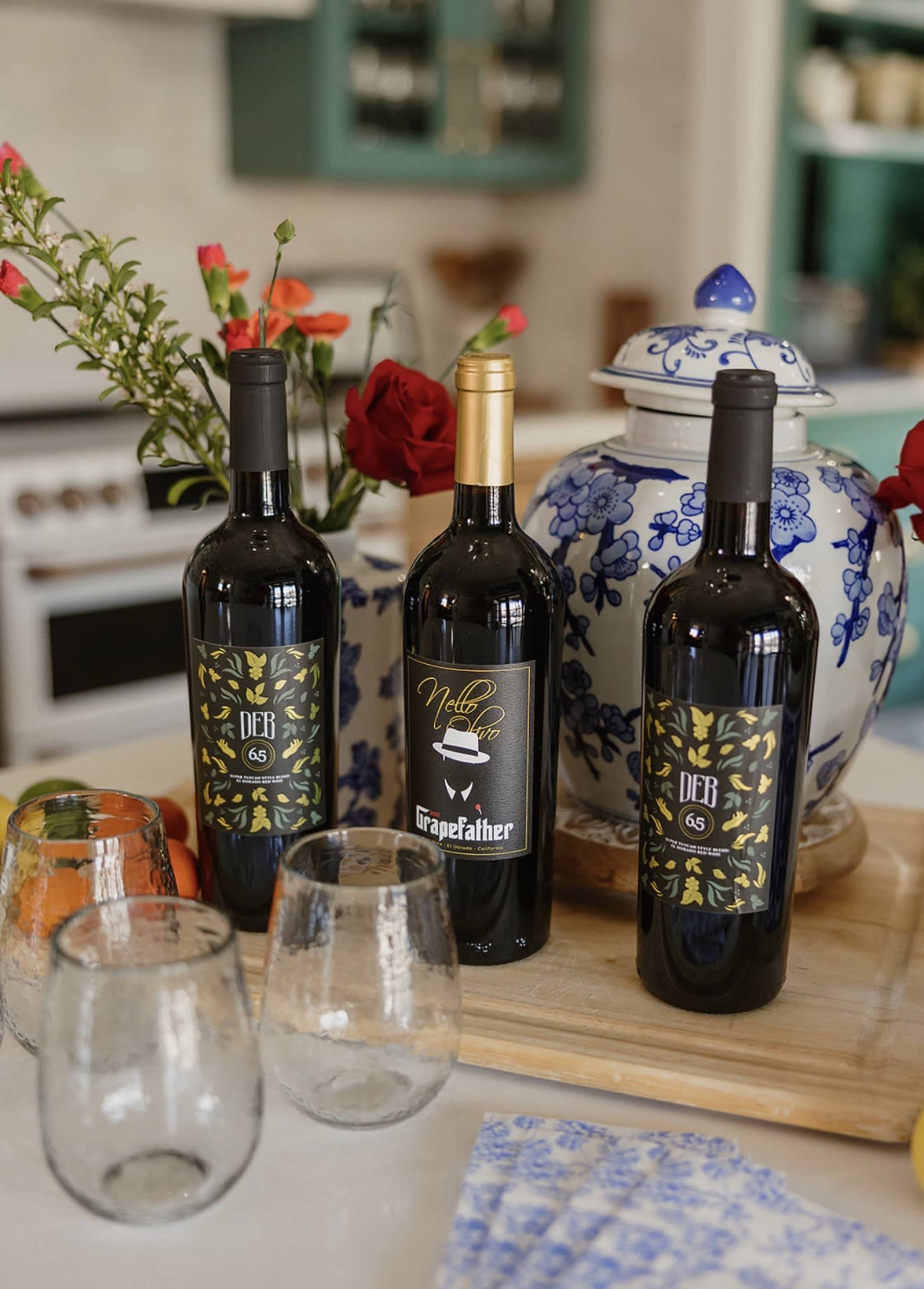 The image features three bottles of wine on a wooden tray, accompanied by two empty glasses and a decorative blue and white porcelain container, with flowers arranged nearby.