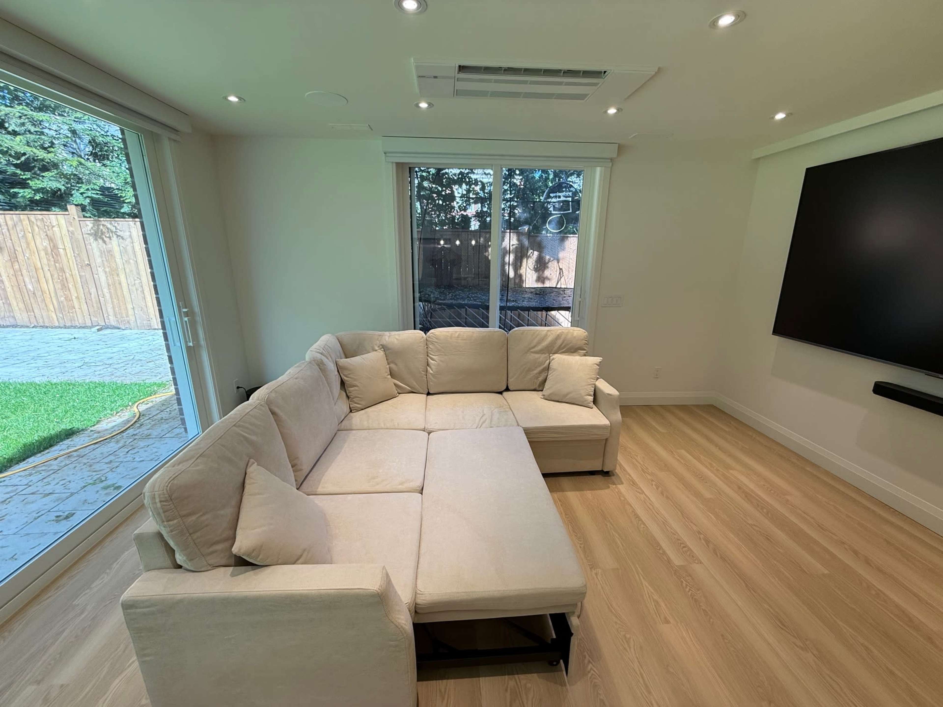 The image shows a light-colored sectional sofa positioned in a well-lit room with large windows and a flat-screen television mounted on the wall.