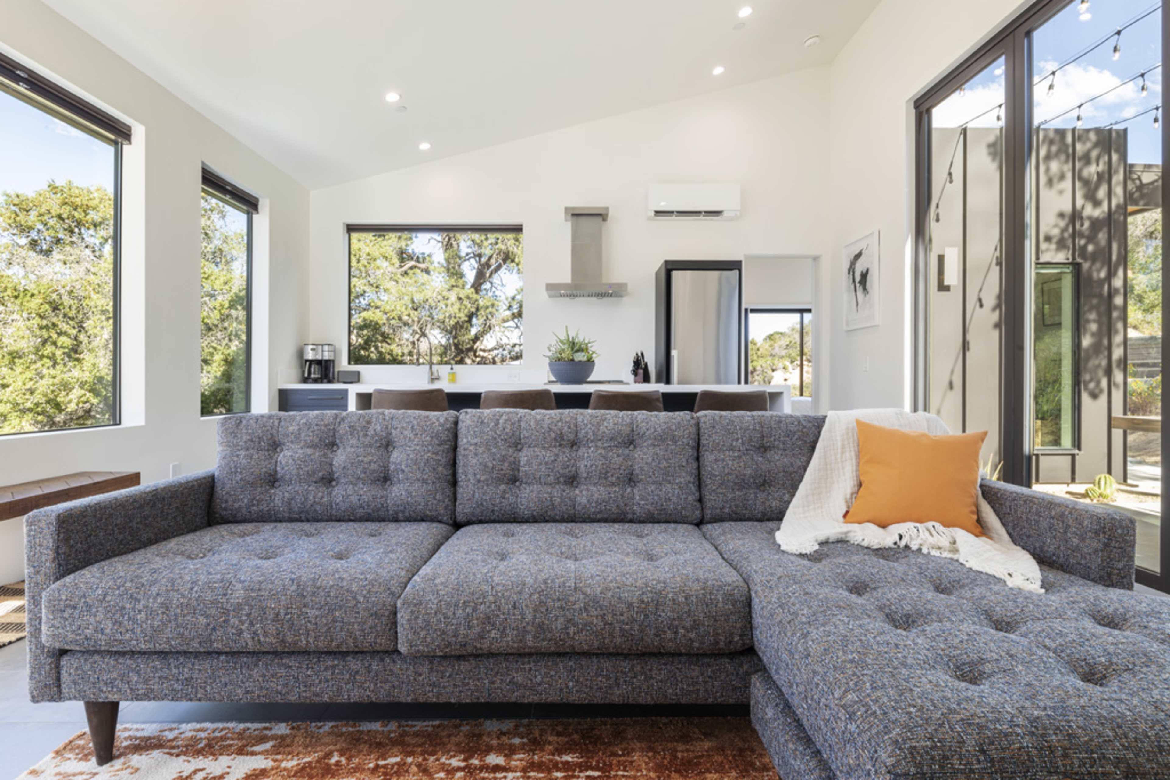 A modern living room features a gray tufted sofa, a wooden bench, and large windows with views of greenery, complemented by a kitchen area in the background.