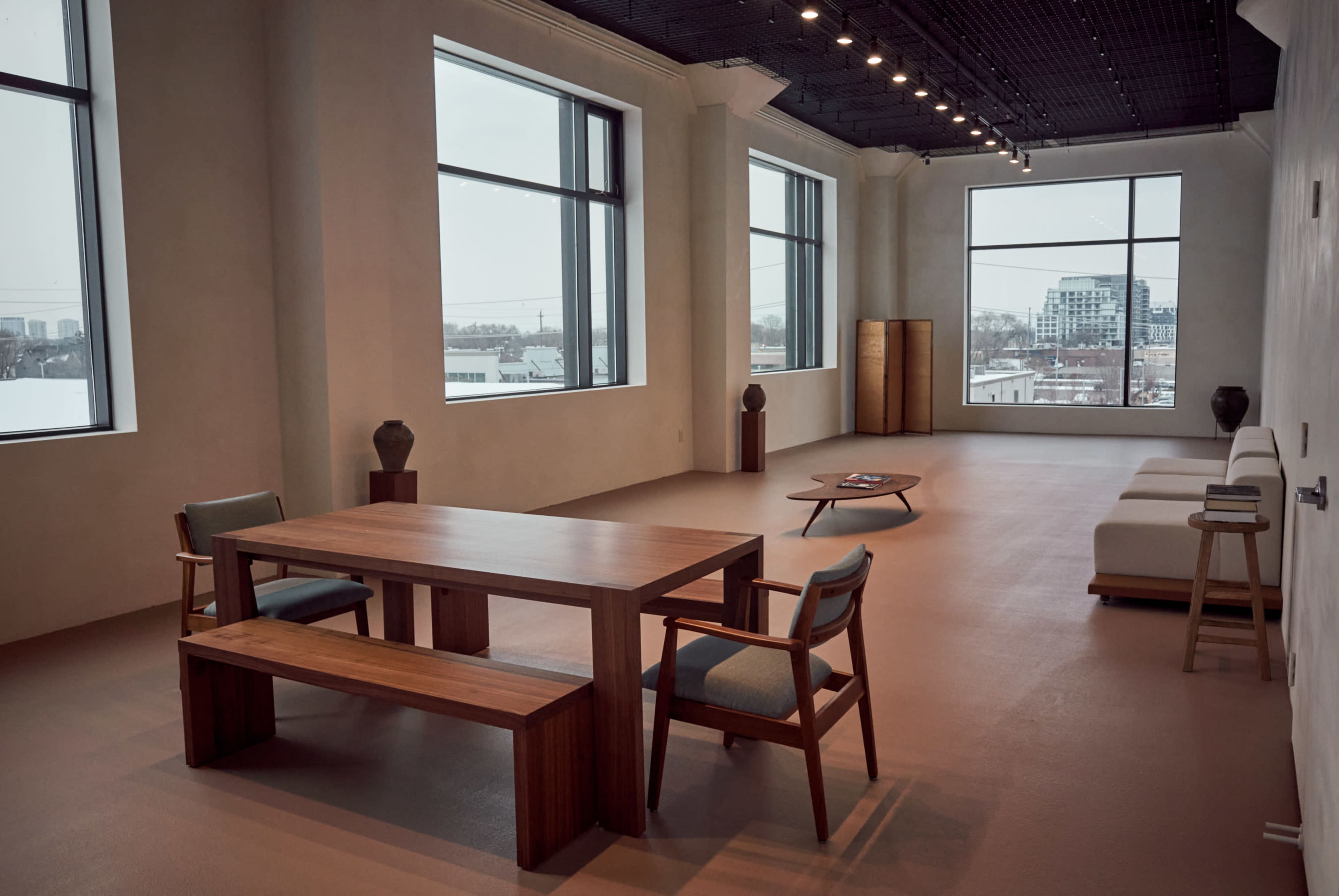 The image shows a spacious, minimally furnished room with large windows, featuring a wooden table, bench, chairs, and a sofa.