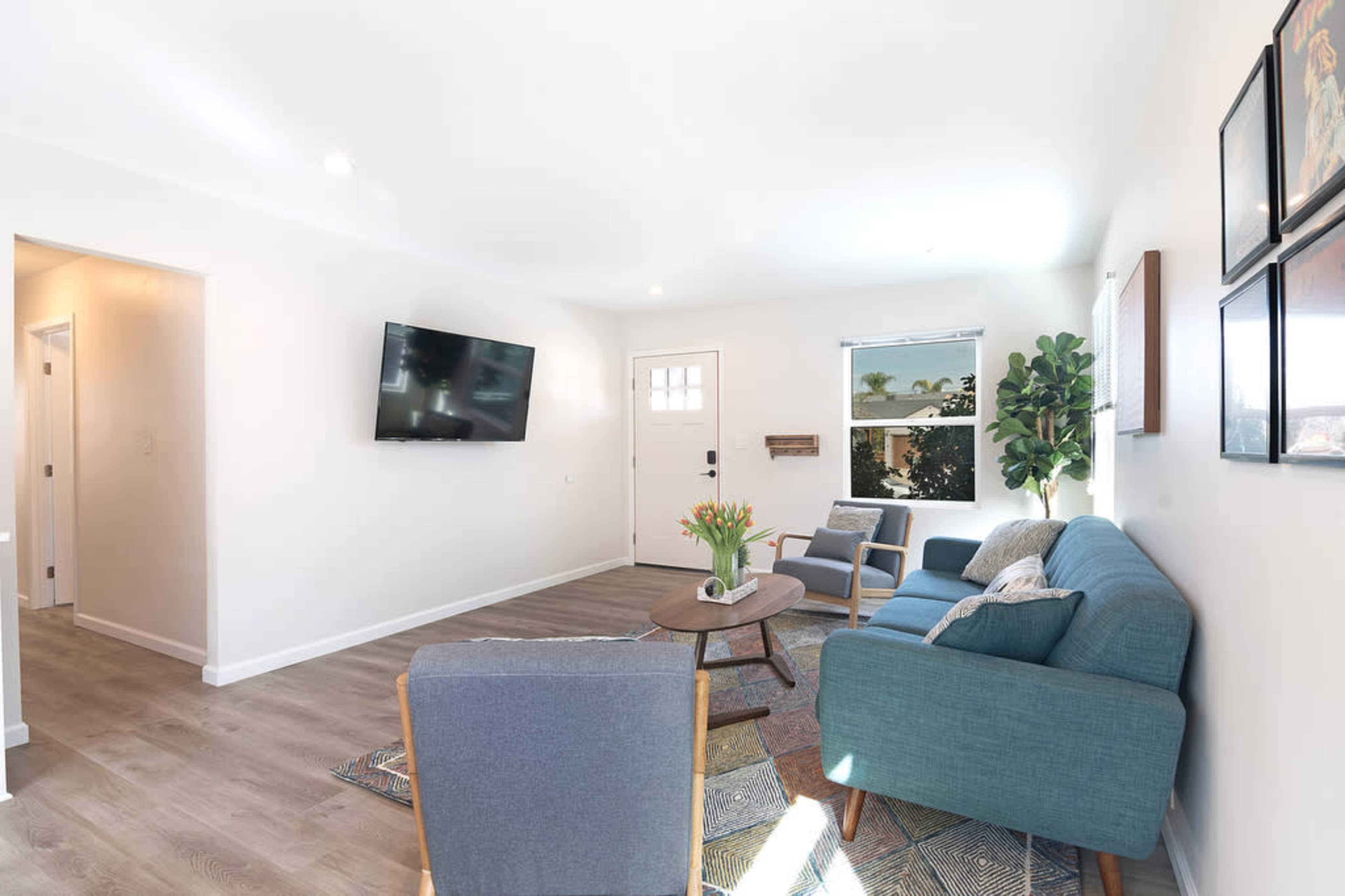 A modern living room featuring a blue couch, a chair, a coffee table, and a wall-mounted TV, with a front door and window allowing natural light.