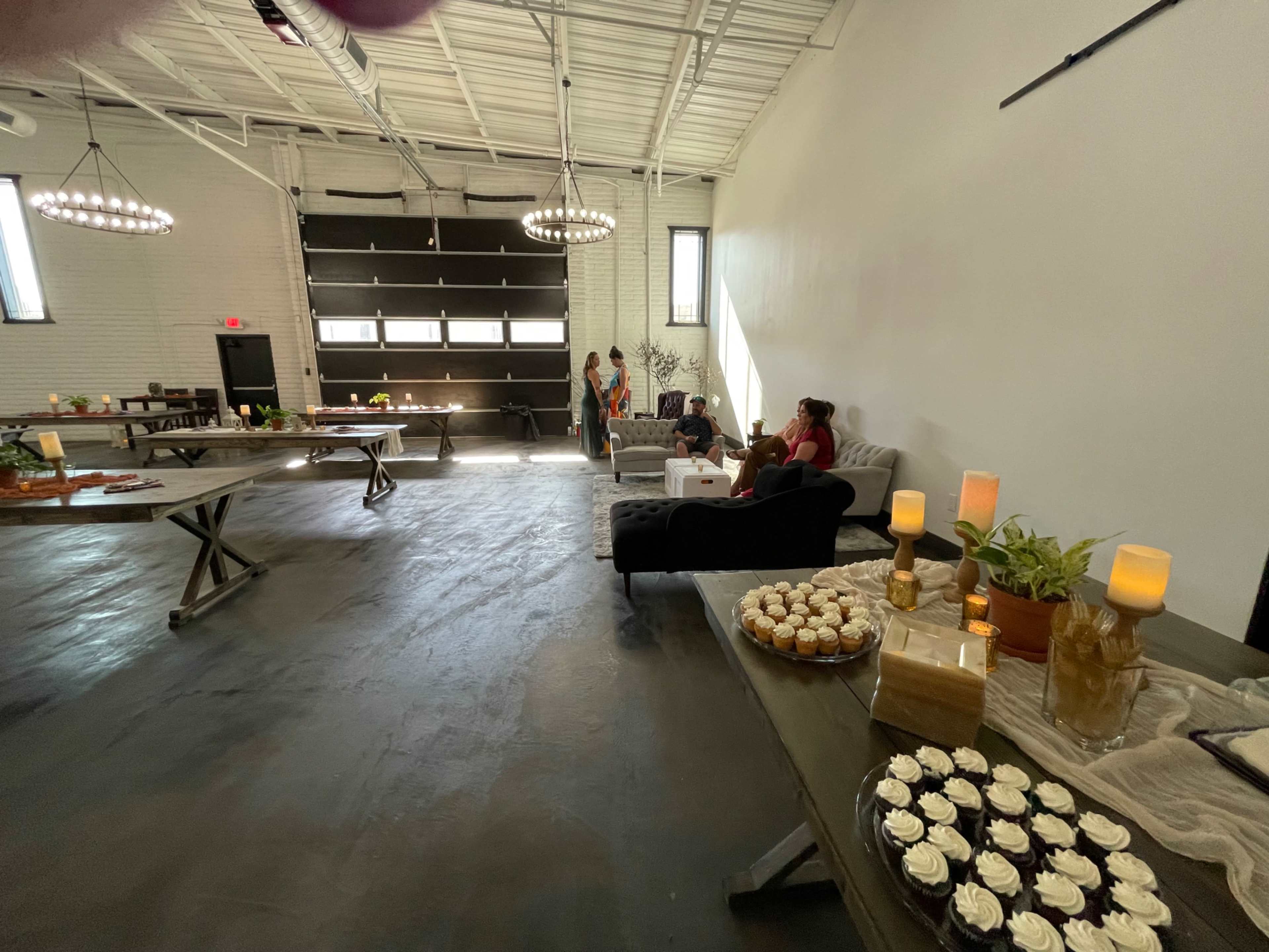 The image shows a spacious indoor venue with tables arranged for an event, featuring desserts on display, and several people seated in a lounge area.