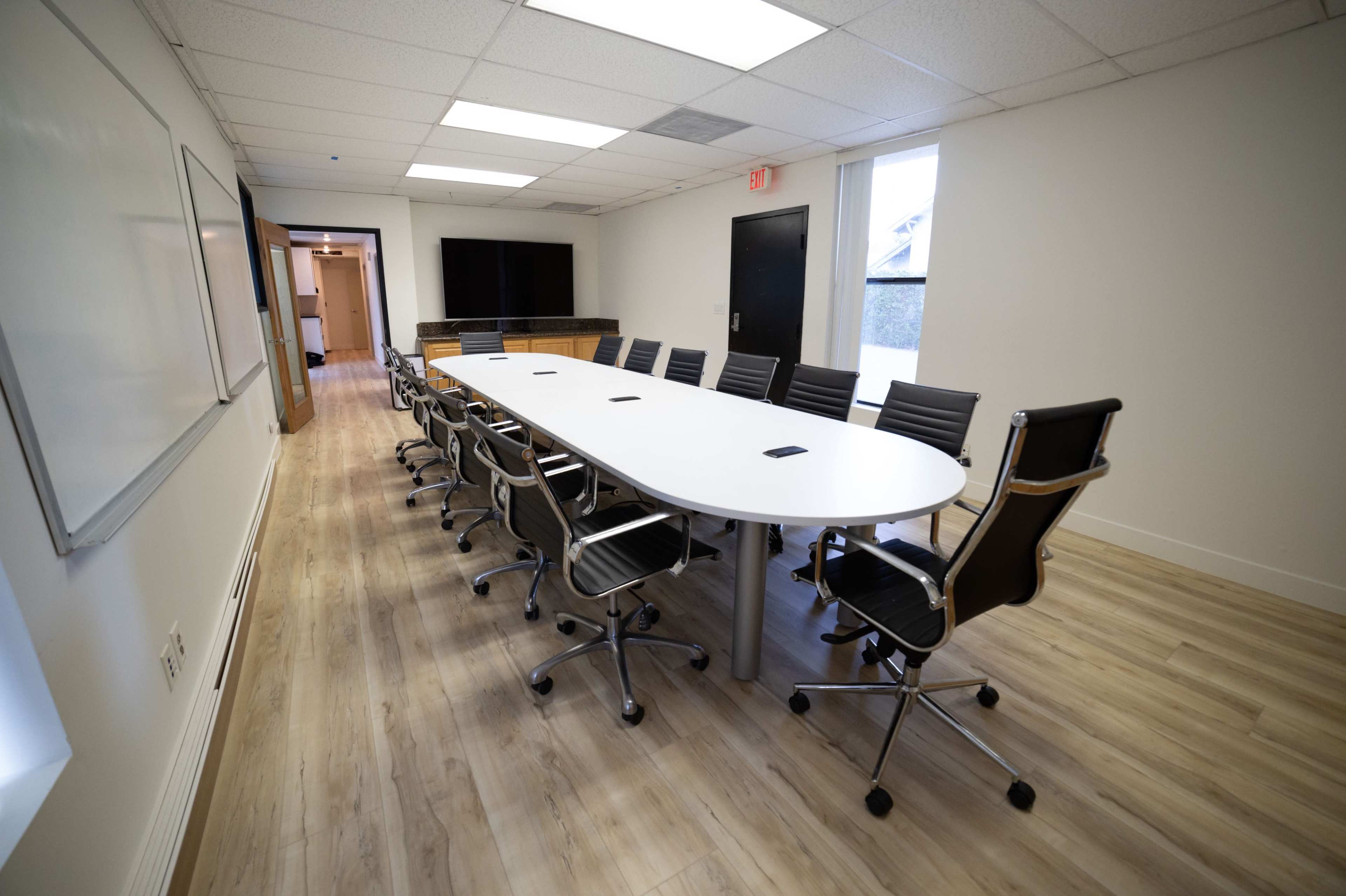 A modern conference room with a long white table surrounded by black office chairs, featuring a whiteboard and a large screen on the walls.