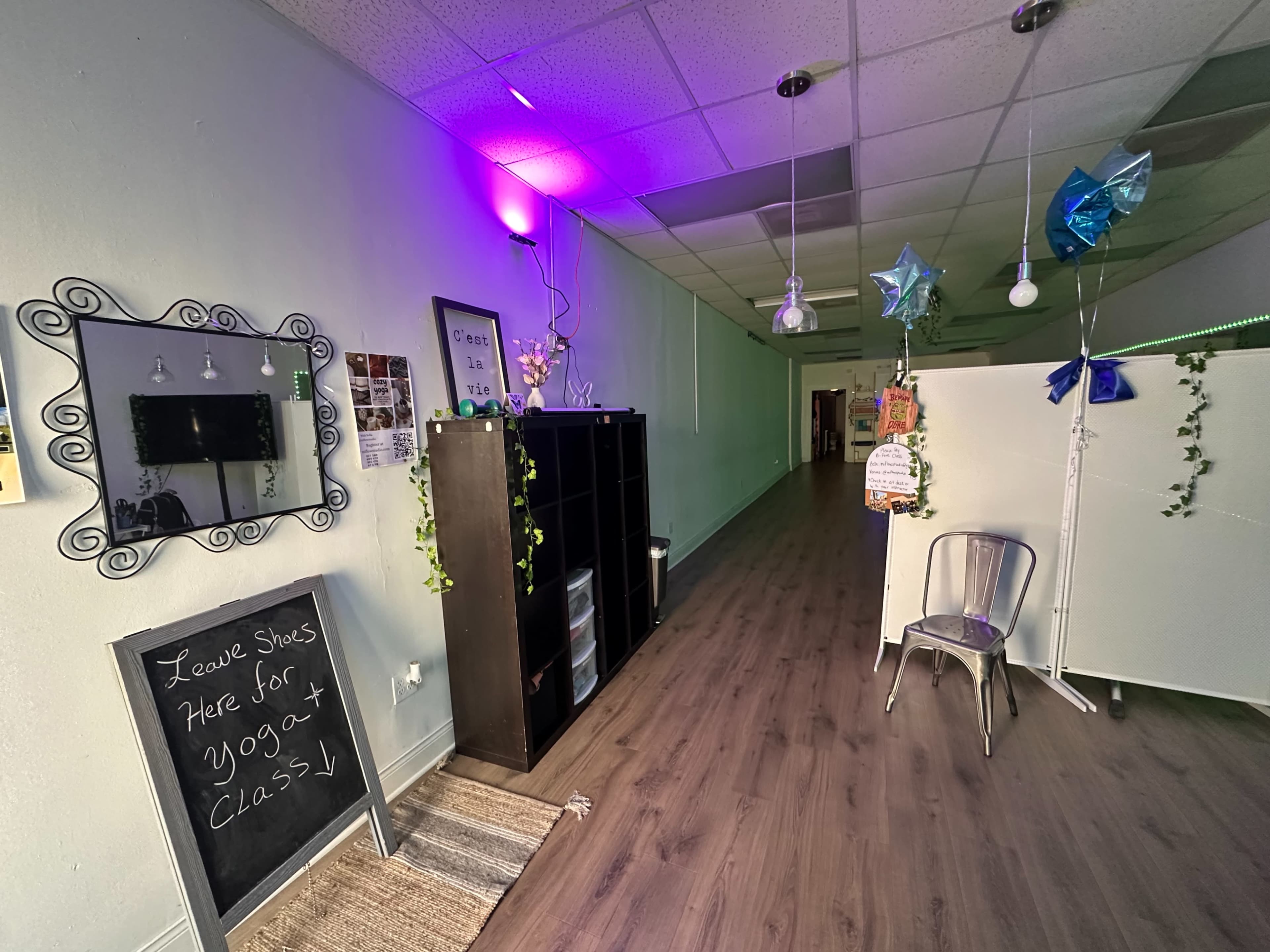 The image shows a hallway of a yoga studio featuring a chalkboard sign, shelving unit, decorative plants, and colorful lighting.