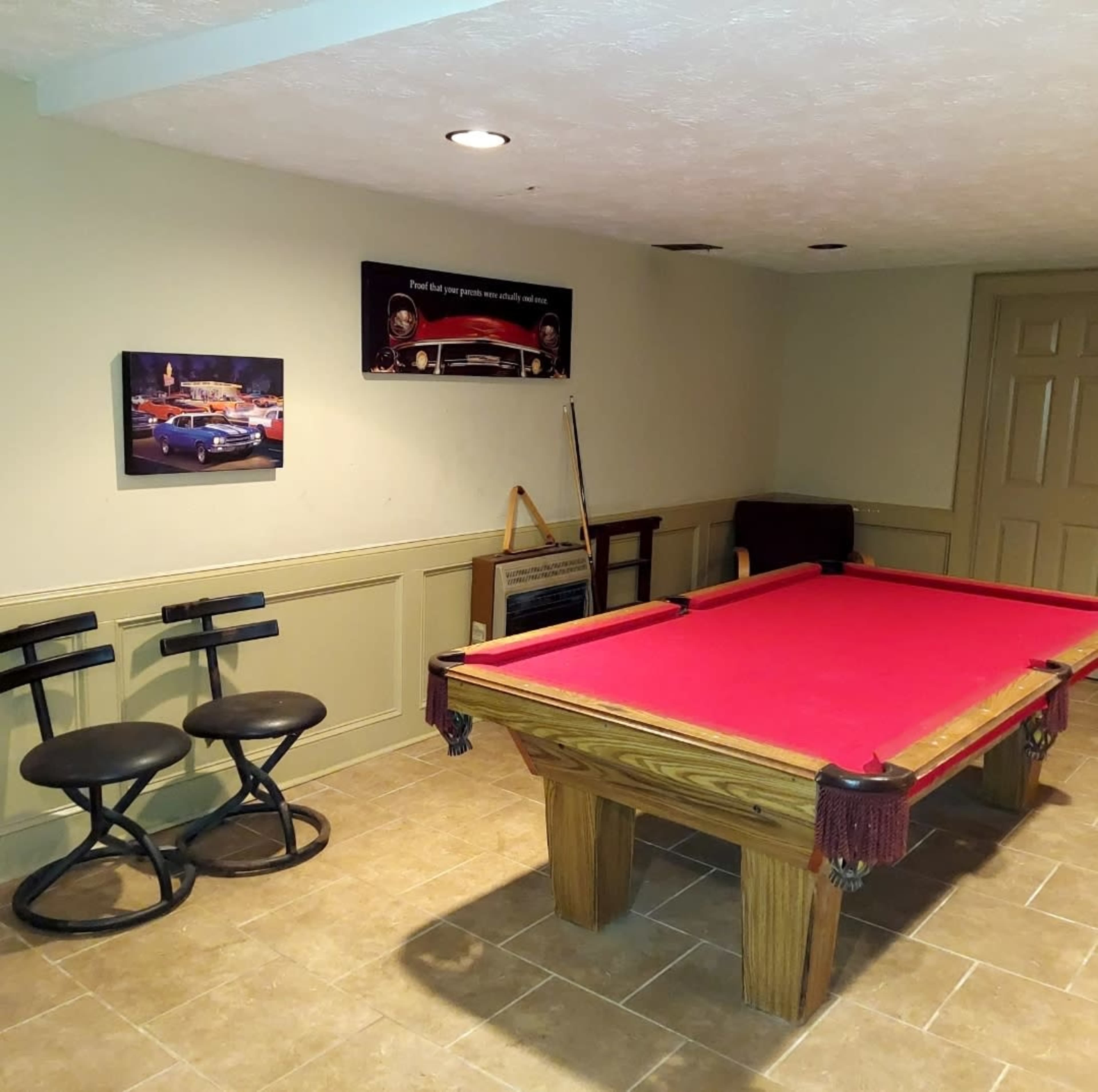 A pool table with a red felt surface, two black stools nearby, and artwork on the wall depicting classic cars.