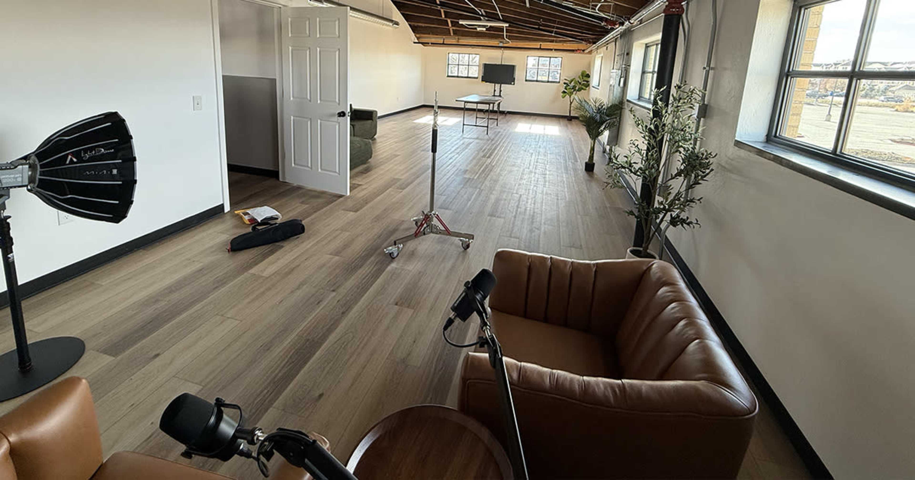 The image shows a spacious room with wooden flooring, featuring a sofa, a coffee table, and photography equipment, along with large windows and a doorway.