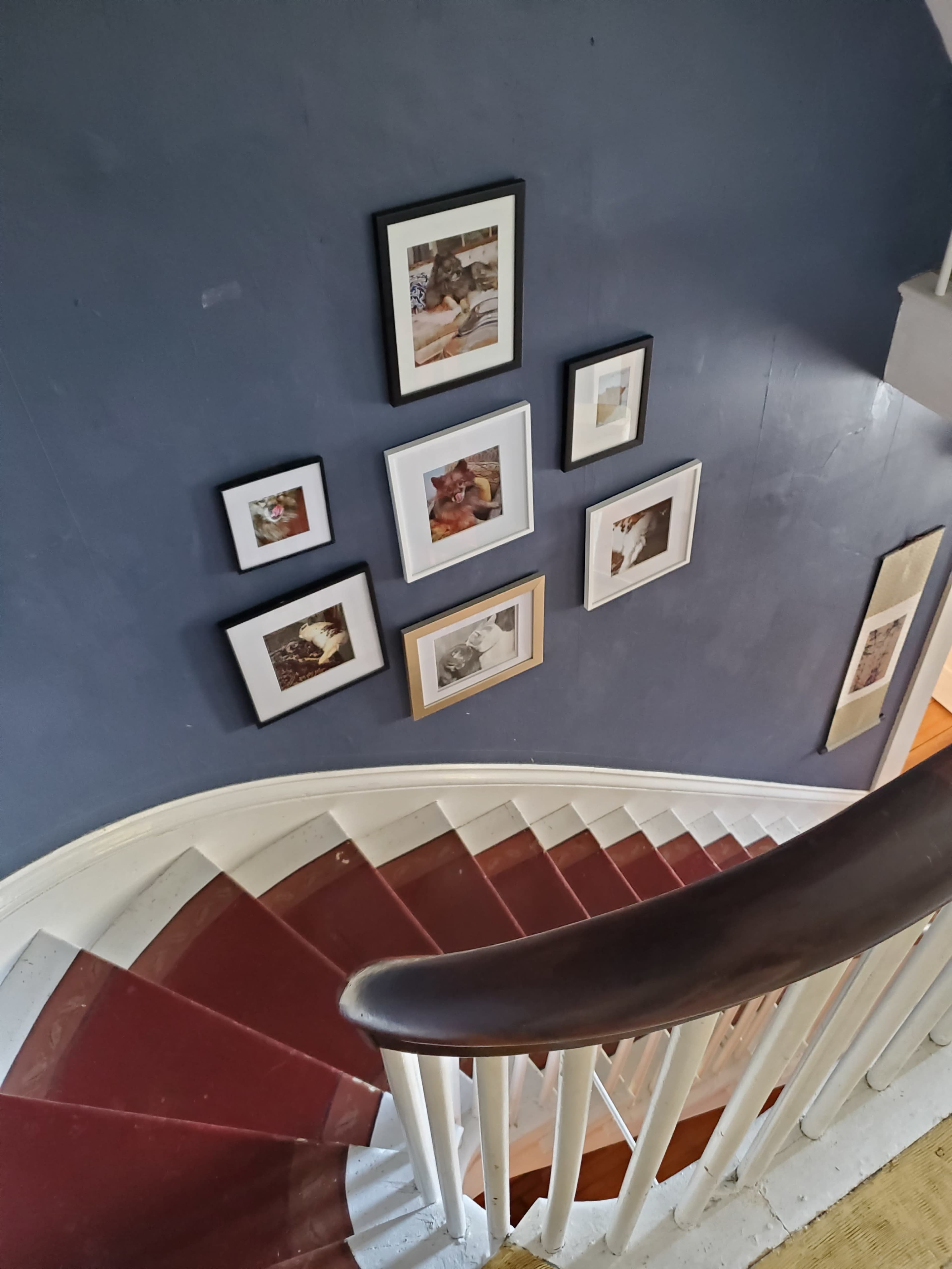 The image shows a spiral staircase with a dark red carpet and a wall adorned with several framed pictures.