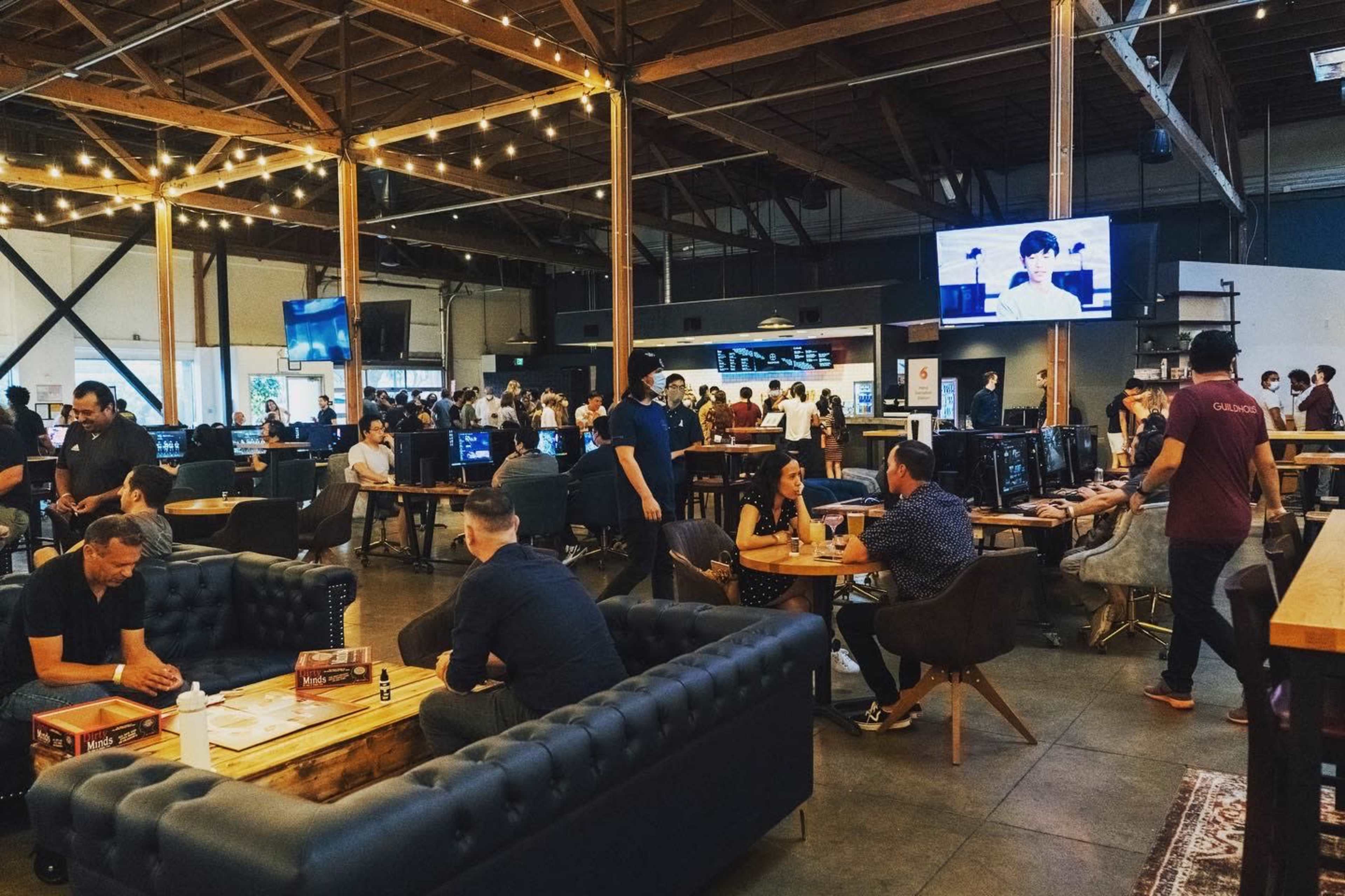 A spacious gaming venue features various groups of people engaged in activities at tables and computers, with bright overhead lighting and a large screen displaying a video.