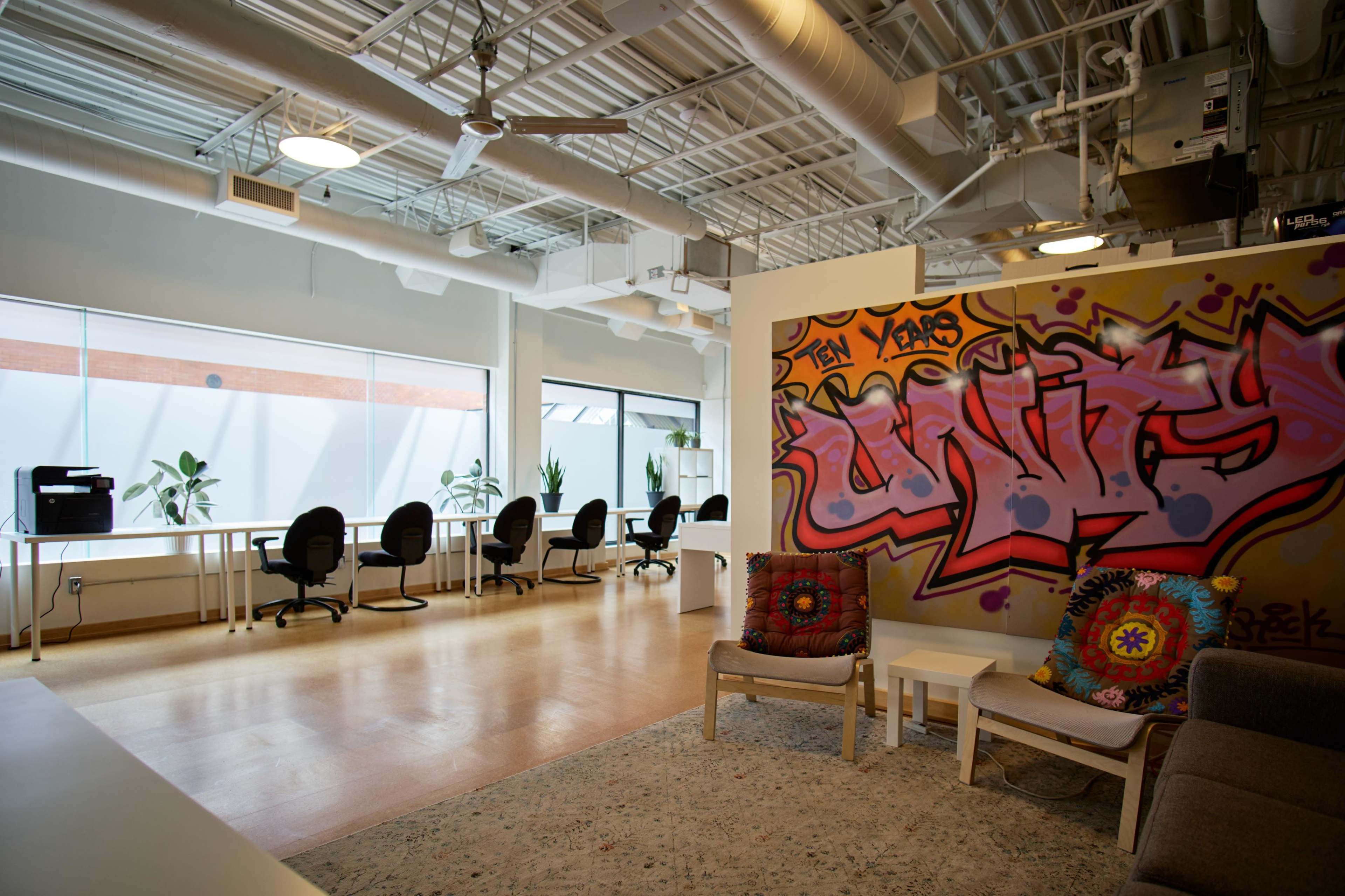 A modern workspace with a colorful graffiti wall, an area for seating, and several desks arranged in a well-lit room with plants and large windows.