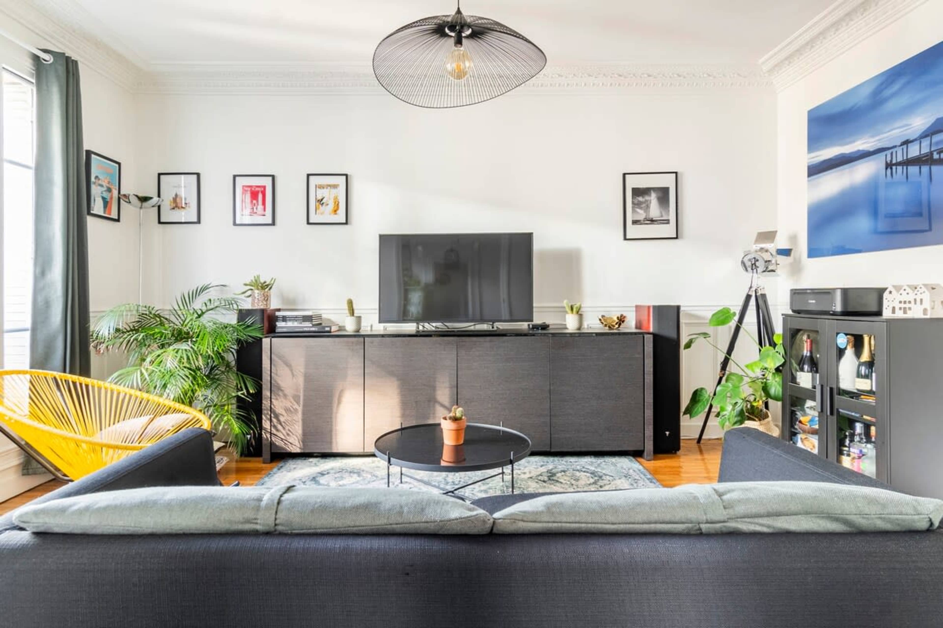 A modern living room with a television on a central cabinet, surrounded by plants and framed artwork on the walls.