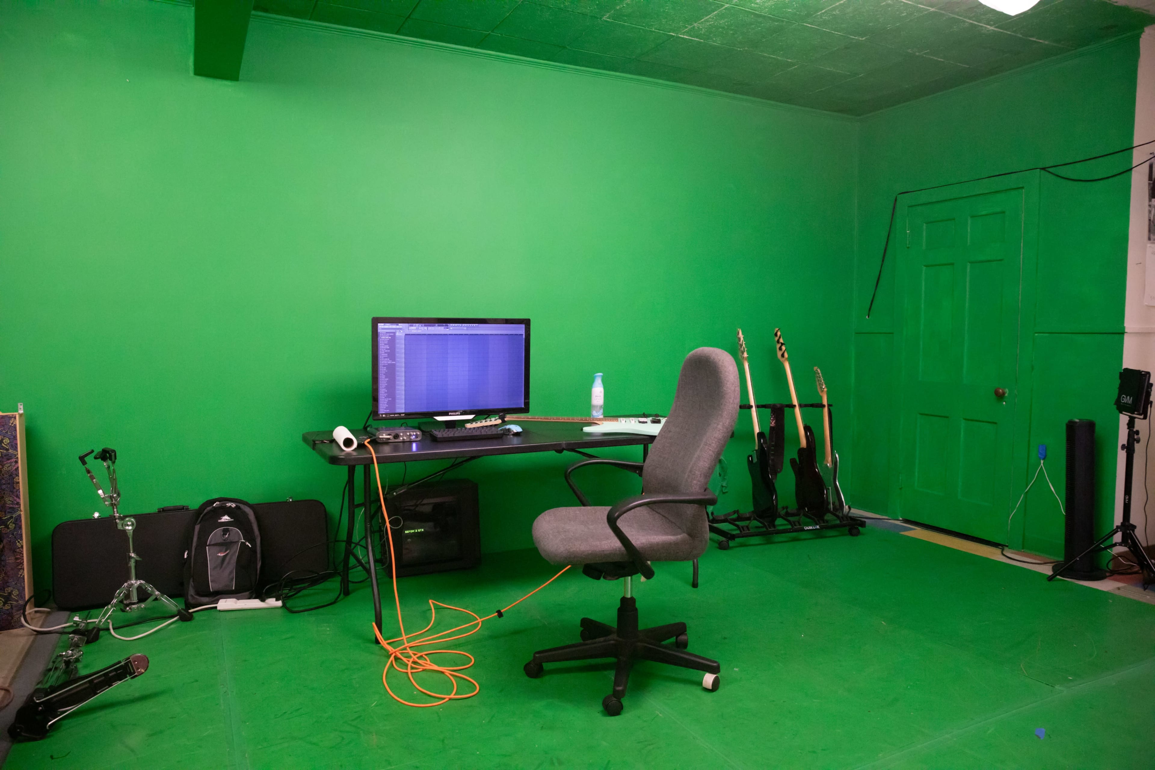 The image shows a green screen room with a chair and computer desk, surrounded by musical instruments and equipment.