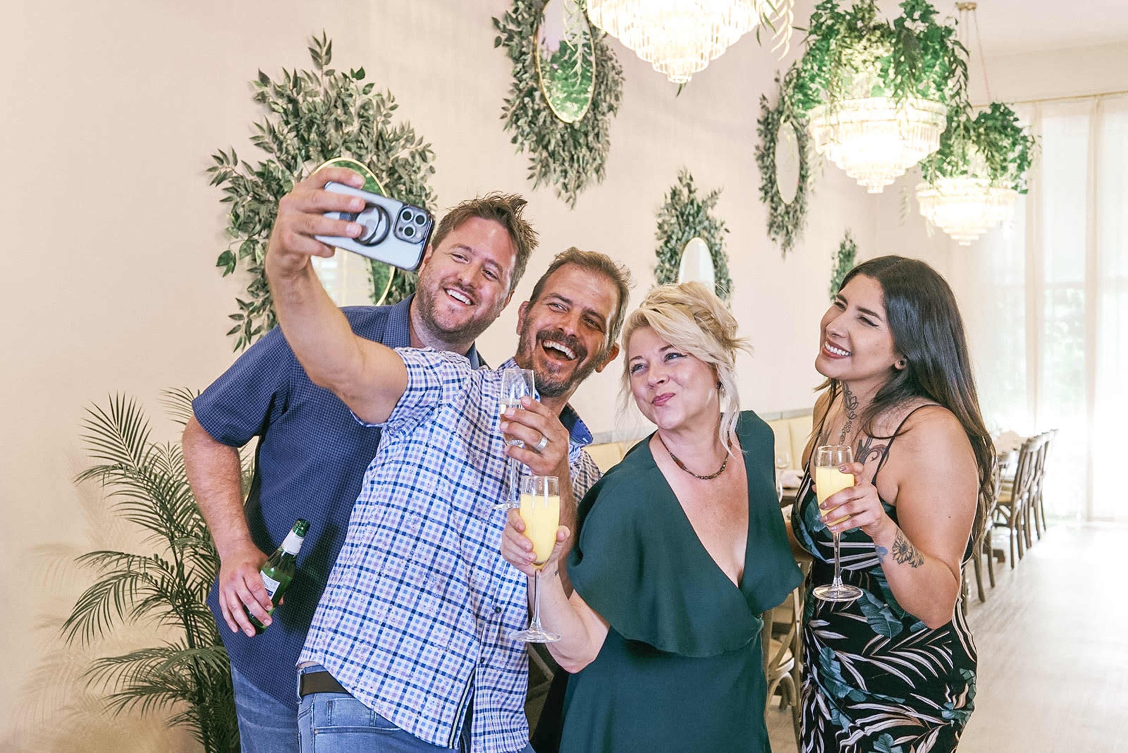 Four people are taking a selfie together while holding drinks in a decorated indoor setting with greenery and chandeliers.