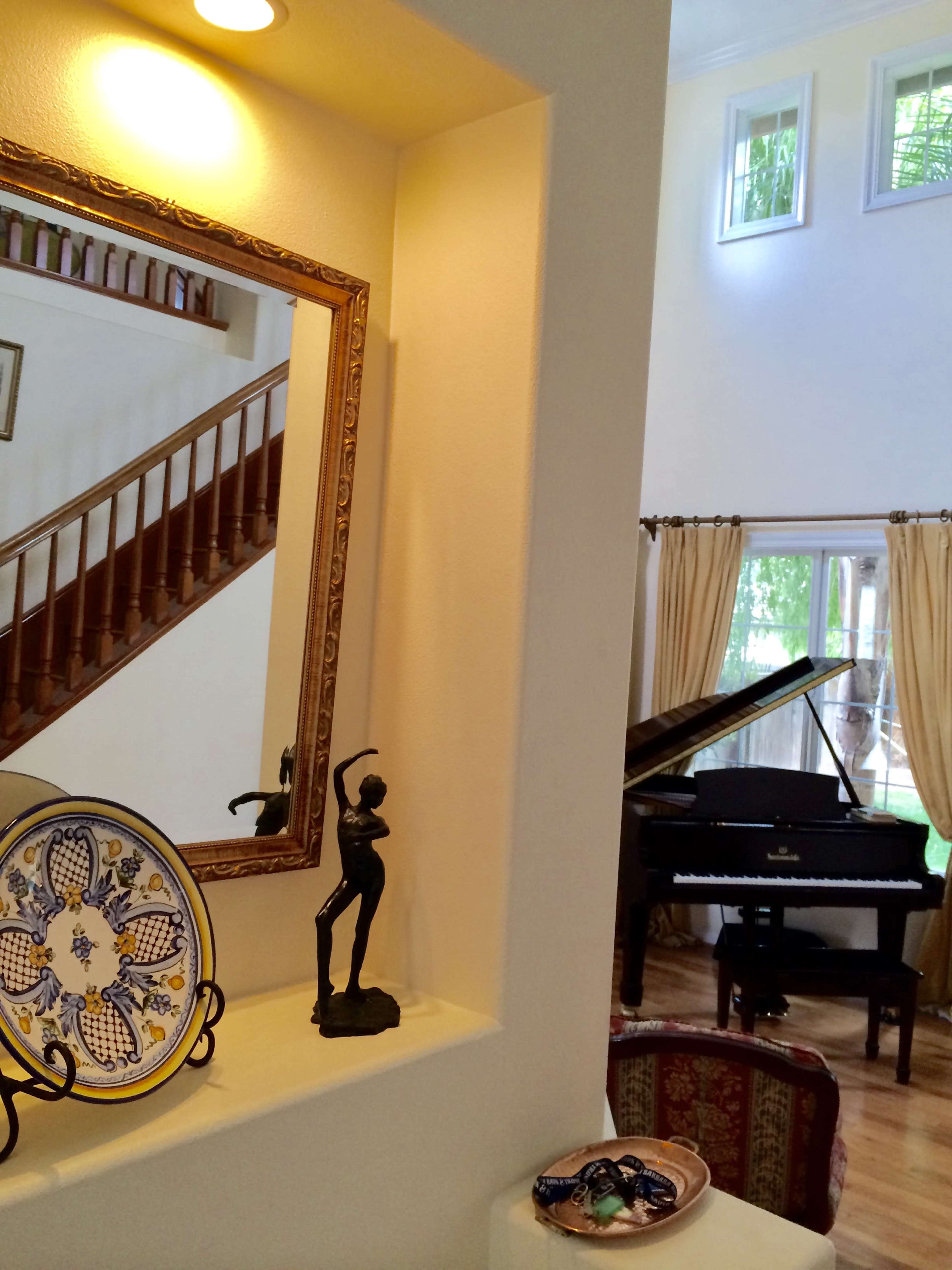 A mirror reflecting a staircase and a grand piano in a well-lit living space adorned with a decorative plate and a small sculpture.