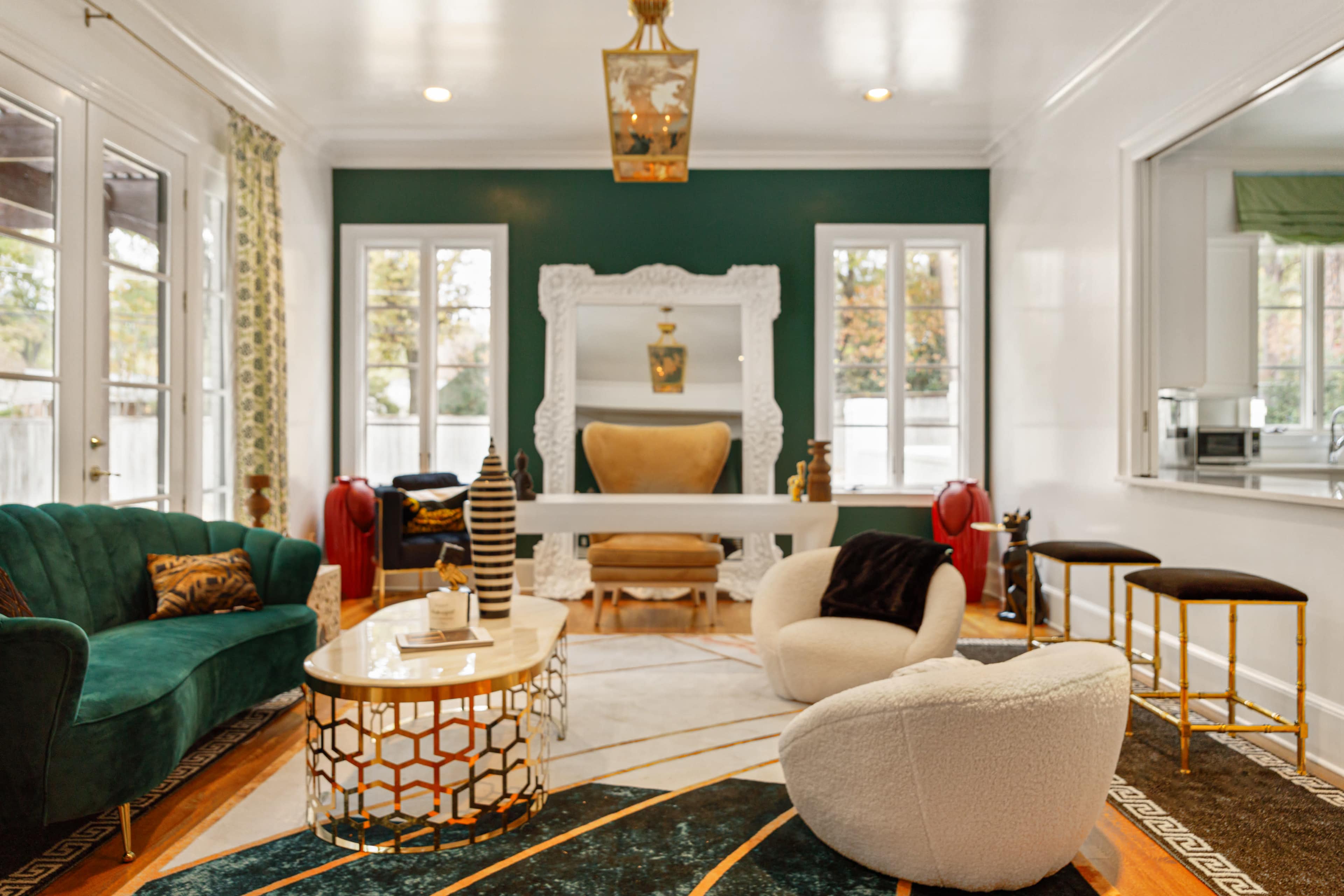 The room features a green accent wall, a large mirror, a mix of seating options, and a coffee table on a patterned rug.