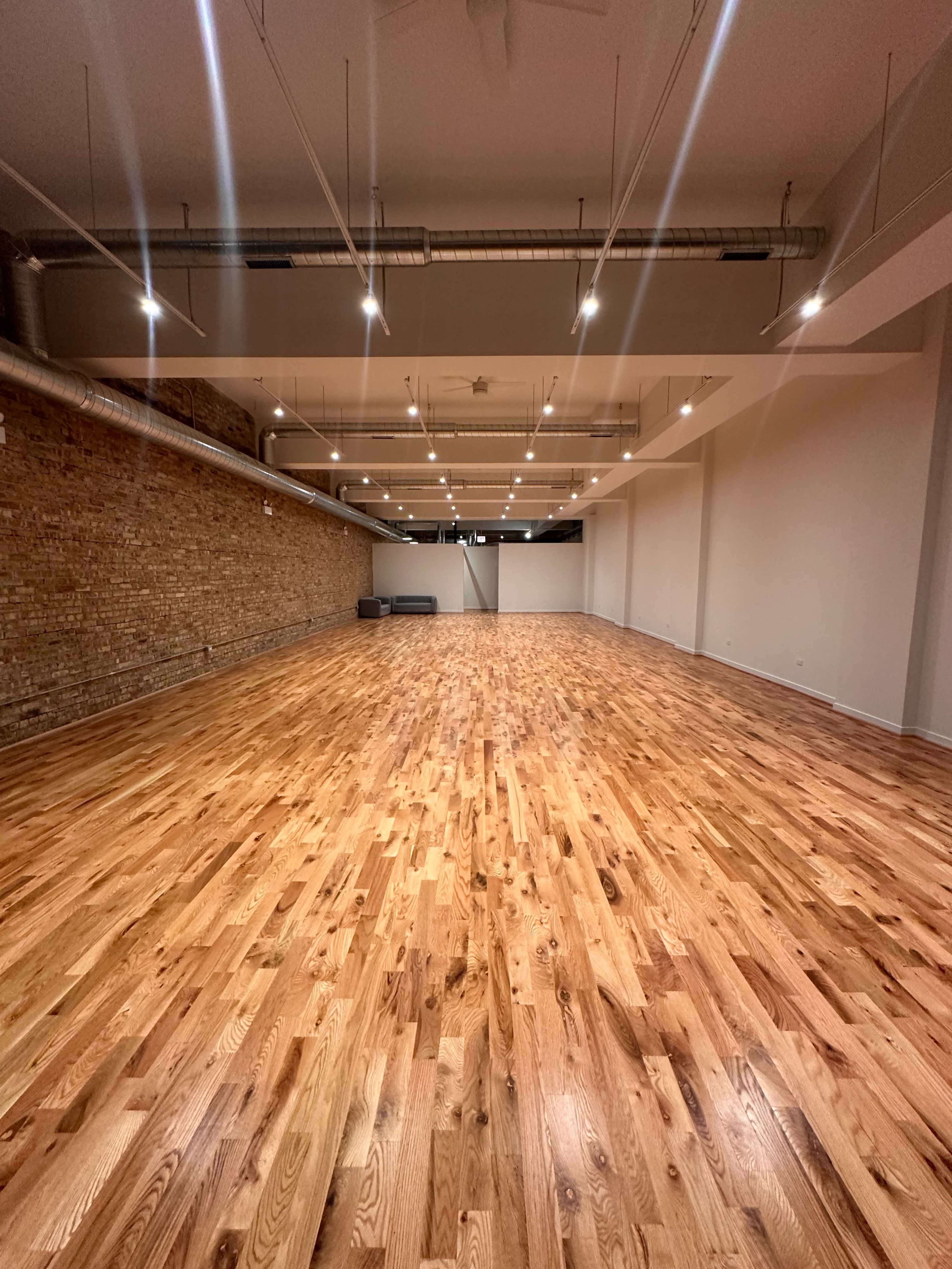 The image shows a spacious, empty room with polished wooden flooring, exposed brick walls, and overhead lighting.
