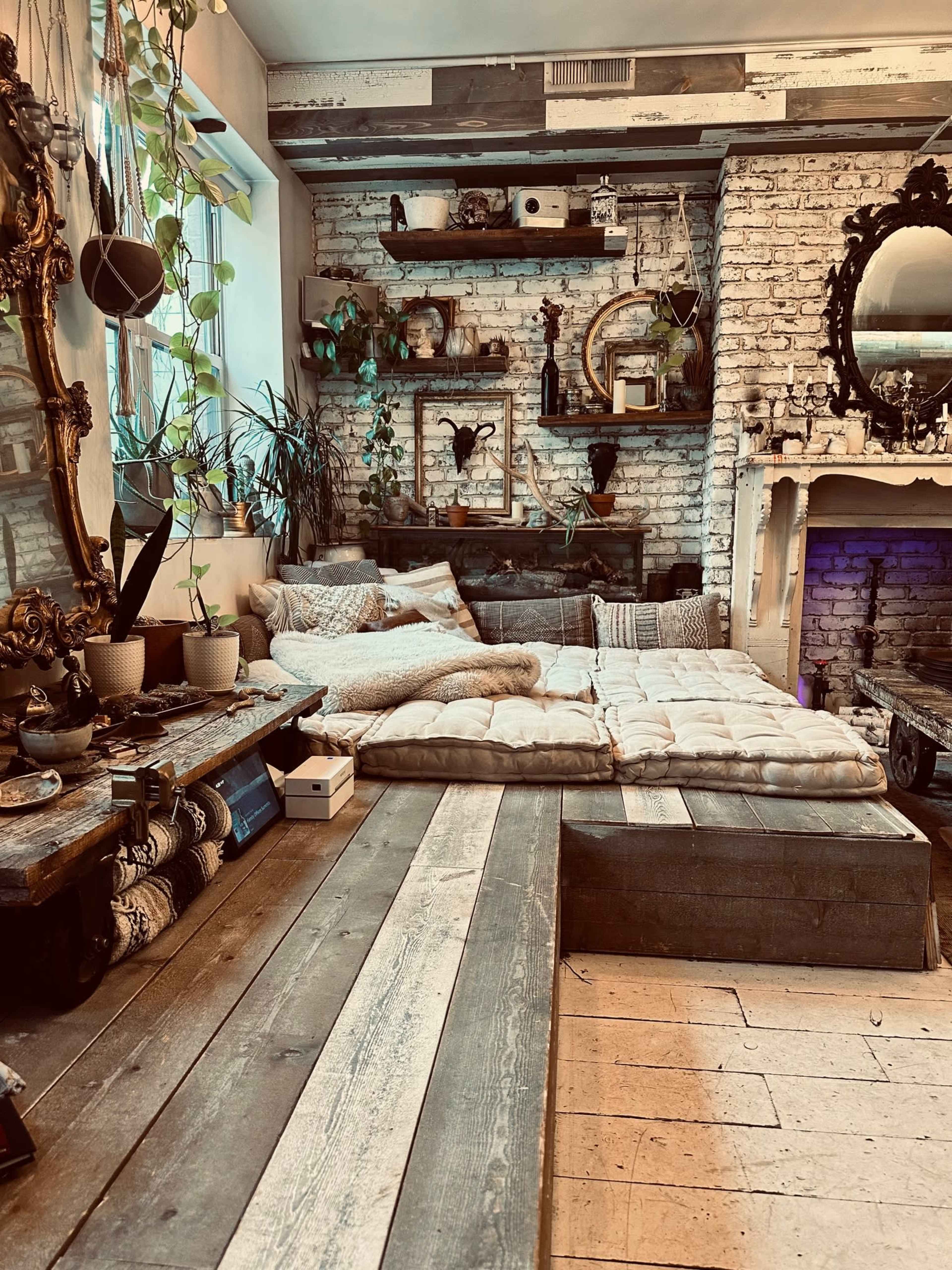 The image shows a cozy indoor living space with a low wooden platform, floor cushions, plants, and a rustic brick wall adorned with shelves and decorative items.