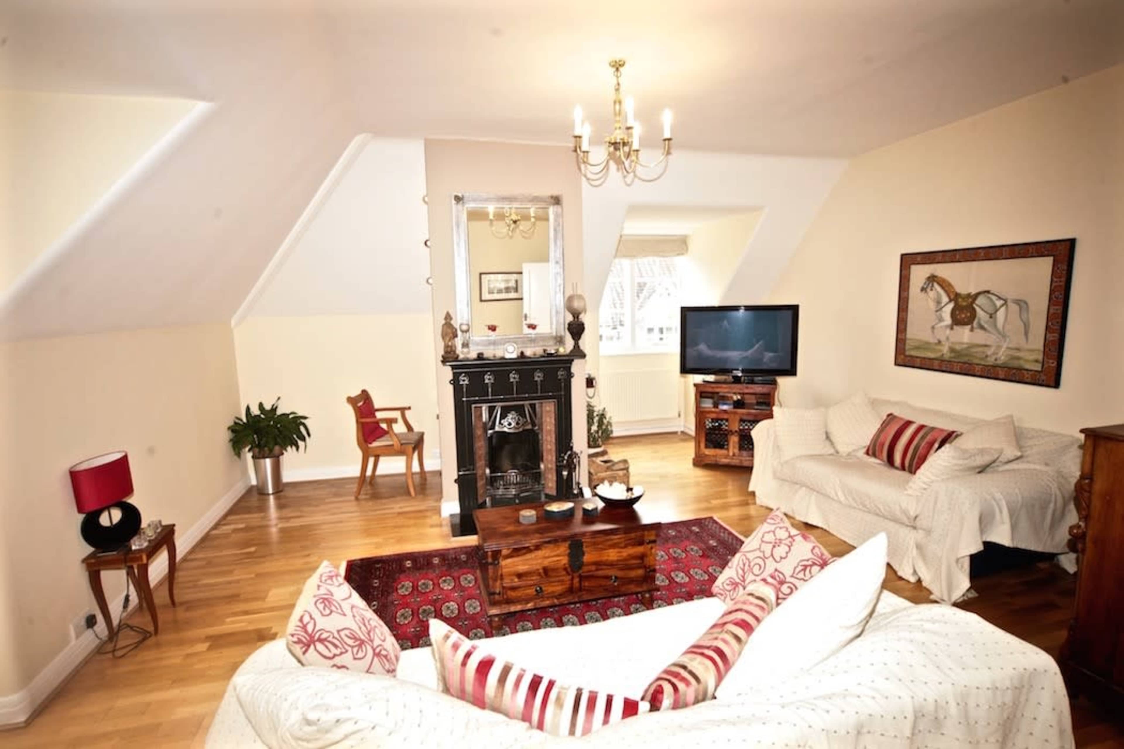 A well-furnished living room with a chandelier, a black fireplace, a television, and seating arranged around a coffee table on a patterned rug.