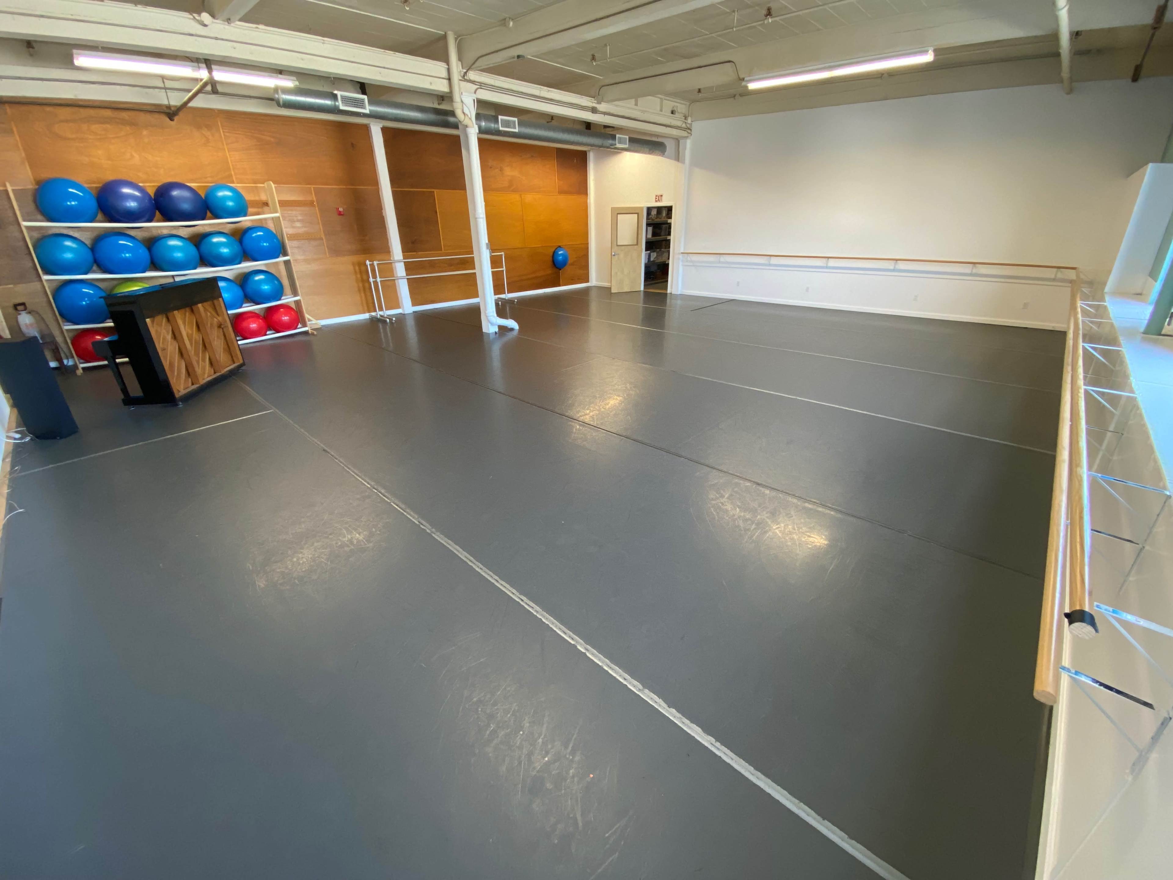 A spacious, empty exercise studio with gray flooring and blue exercise balls stacked against a wooden wall.
