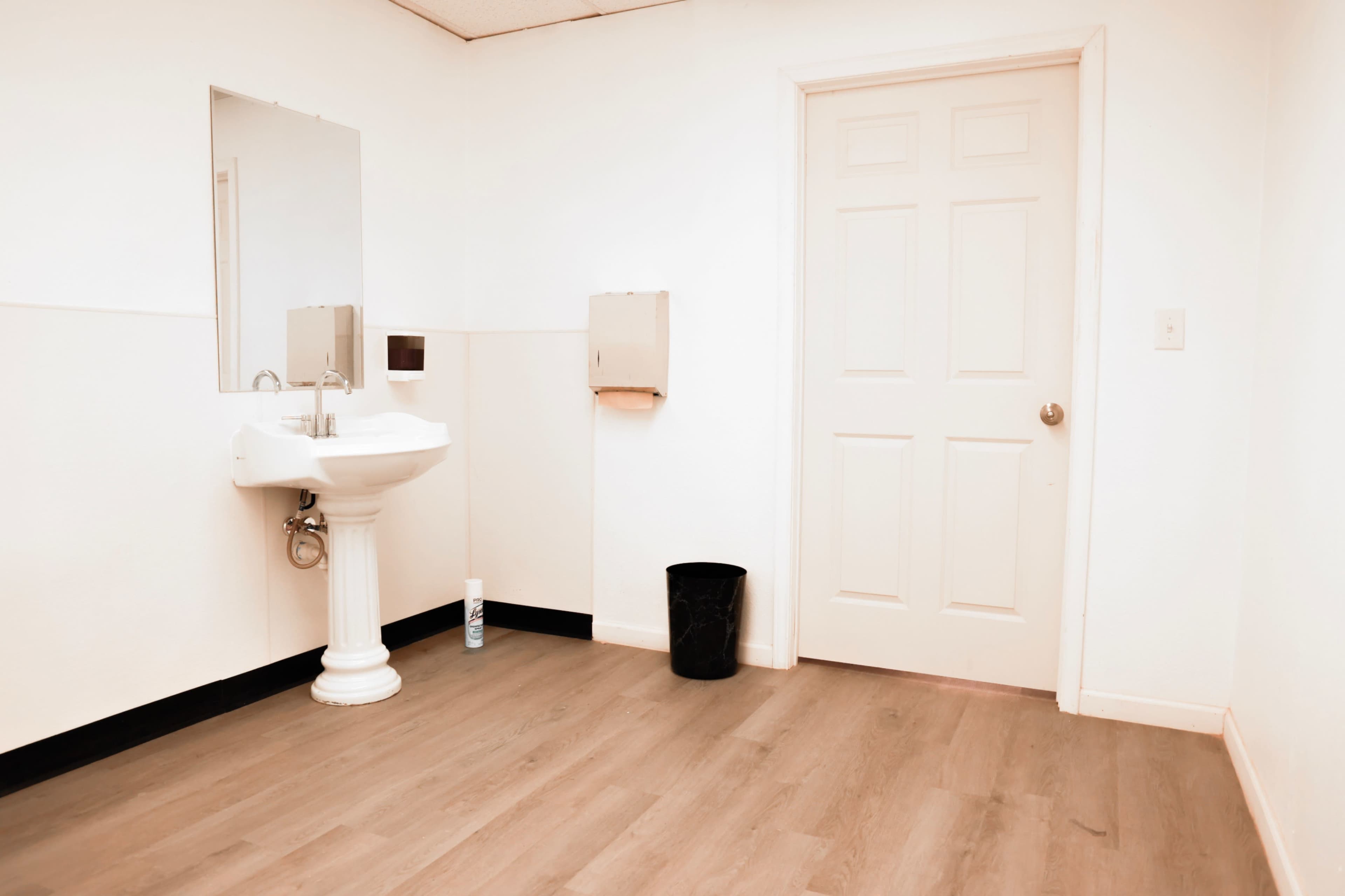 A restroom features a pedestal sink, a mirror, a wall-mounted paper towel dispenser, a trash can, and a closed door, all set against a light-colored wall and wooden floor.