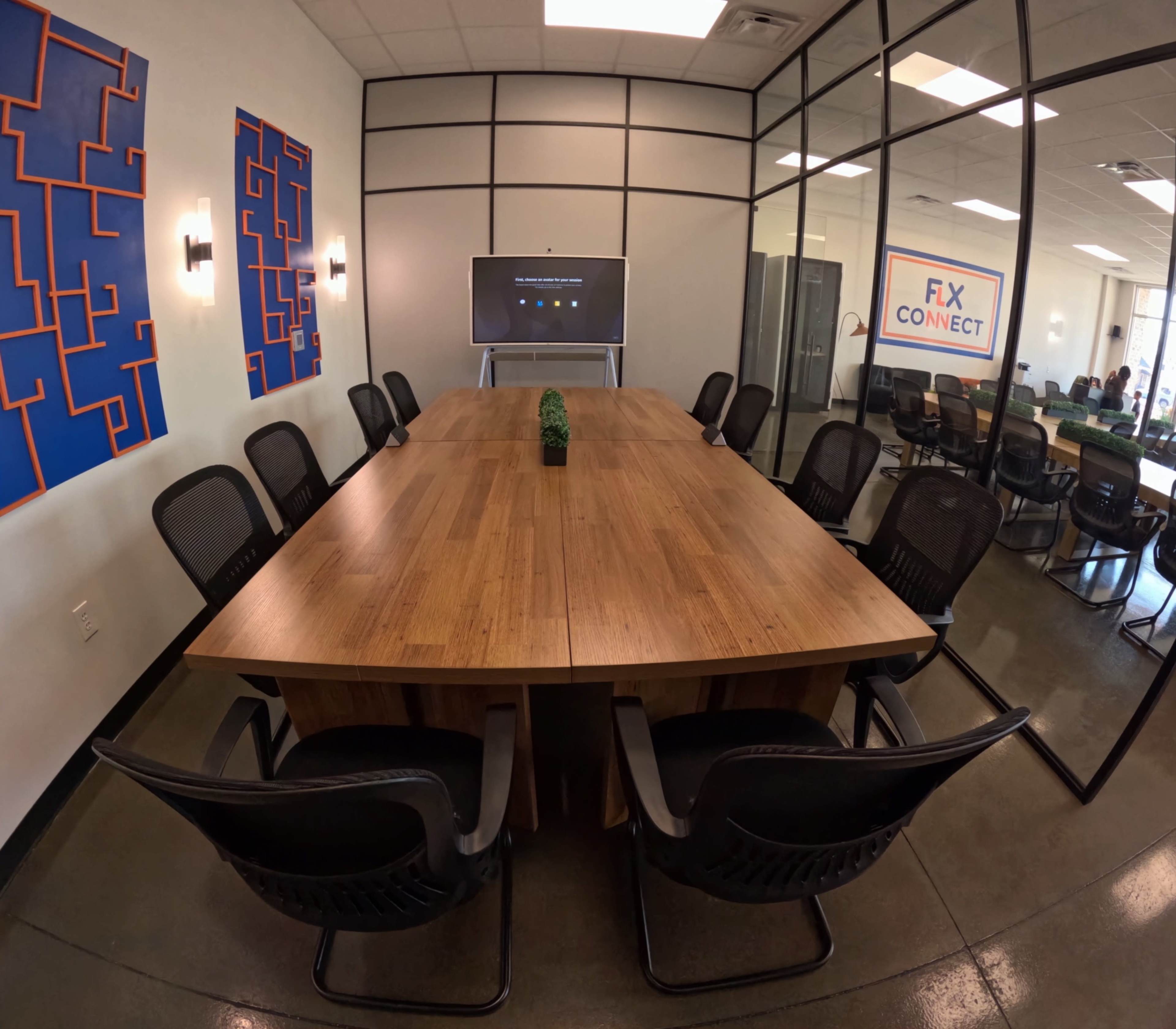 A large wooden conference table surrounded by black chairs is set up in a modern meeting room featuring a wall-mounted screen and abstract wall art.