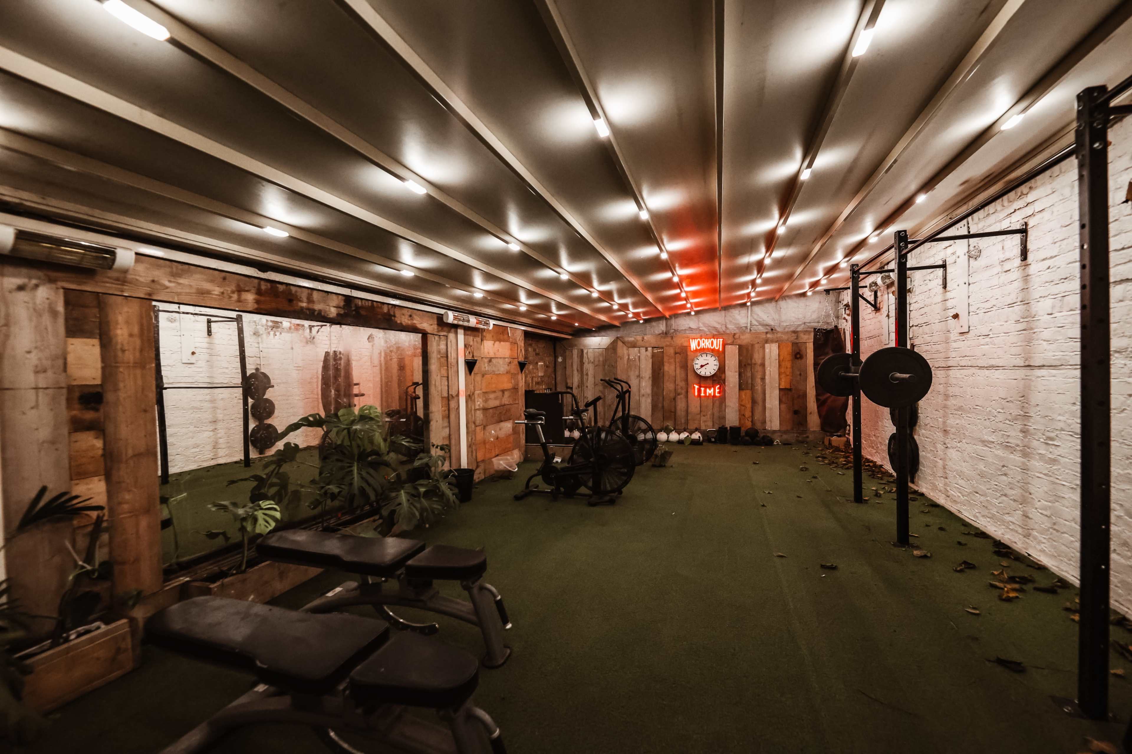 The image depicts a dimly lit gym with exercise equipment, including benches and weights, along with greenery and exposed wooden walls.
