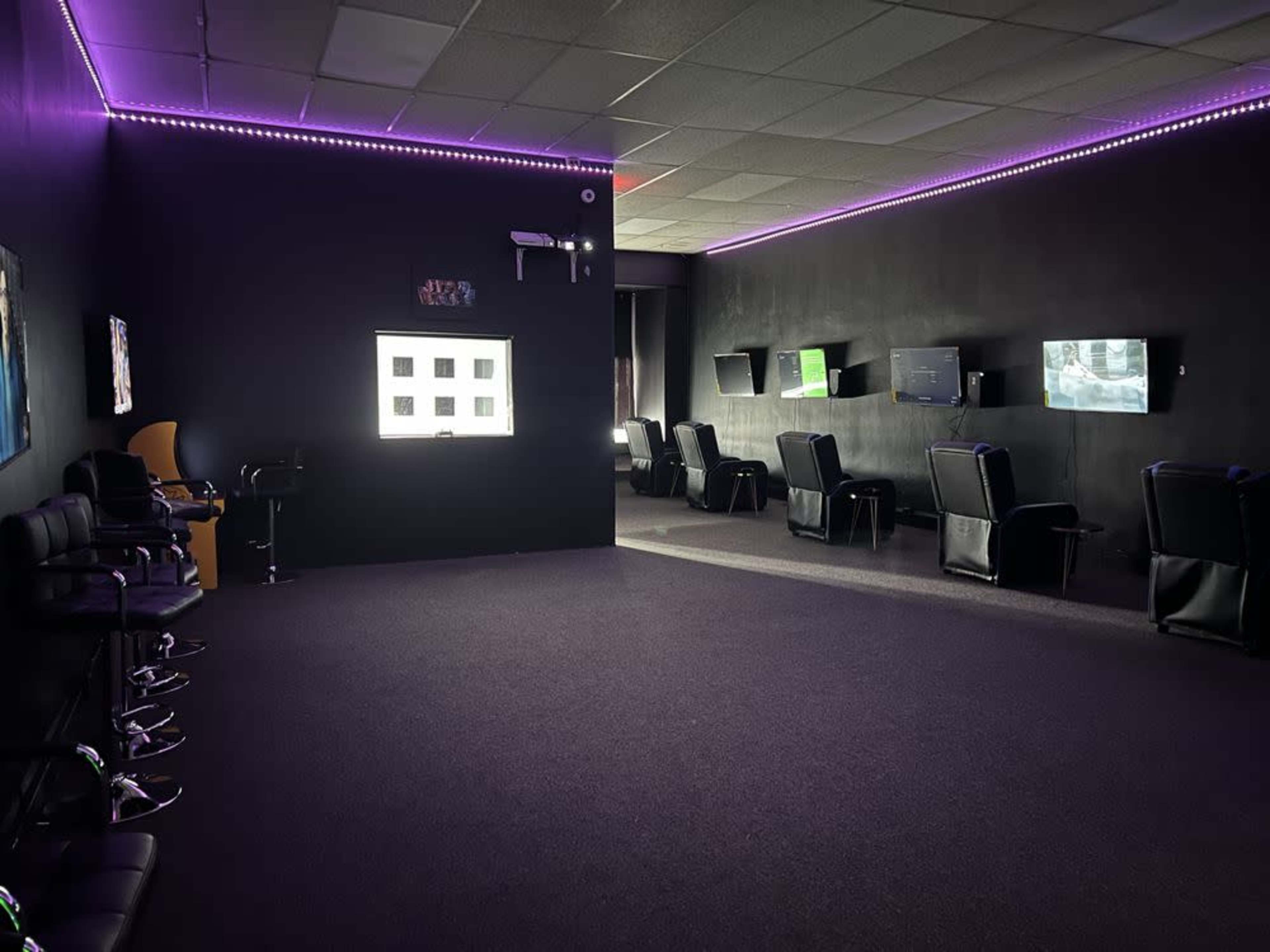The image shows a dimly lit room with black walls, featuring several gaming stations with screens and ergonomic chairs, accented by purple LED lights along the ceiling.