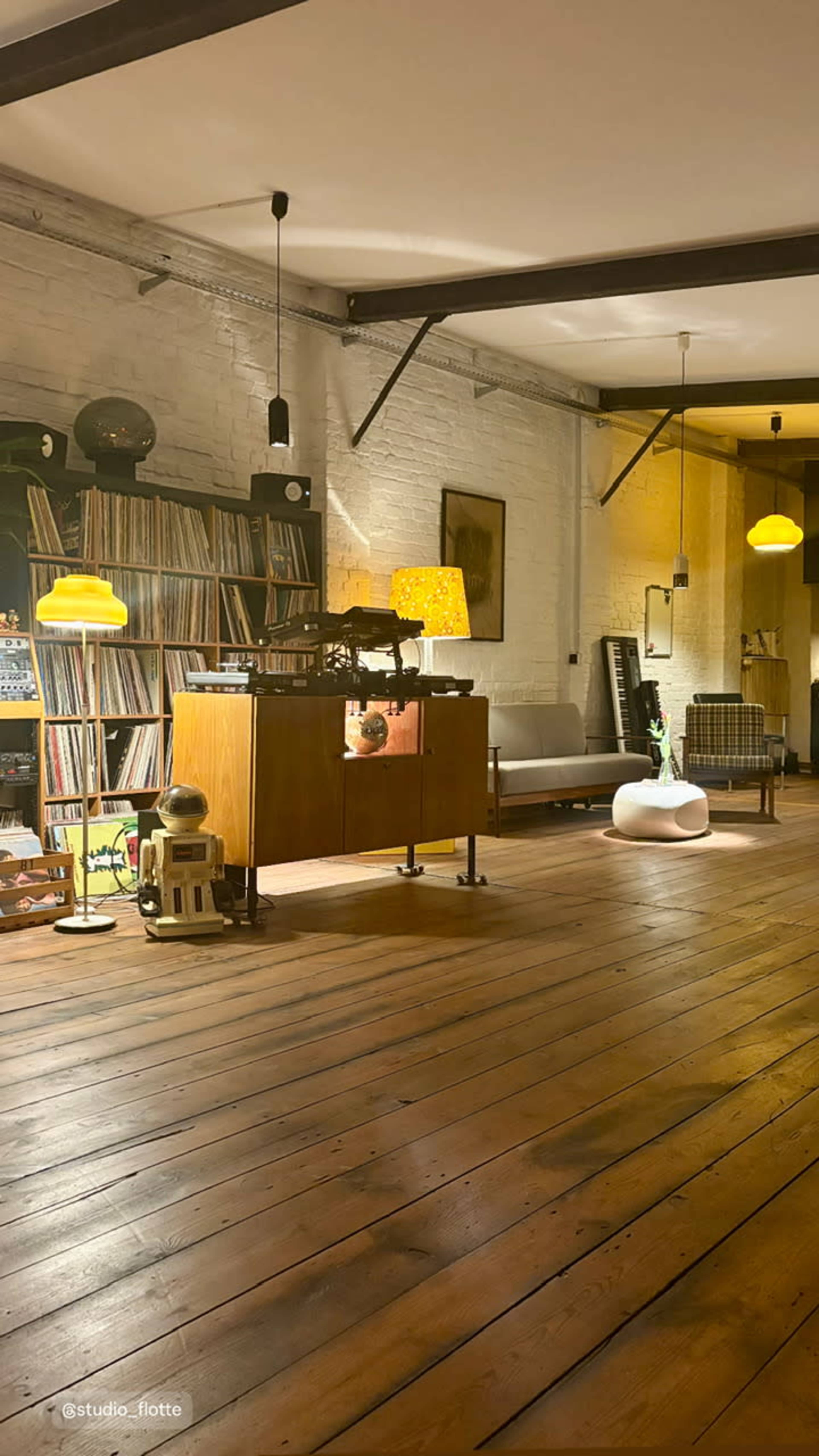 The image shows a spacious, well-lit room with wooden floors, featuring a collection of vinyl records on shelves, a vintage cabinet, and modern furniture.