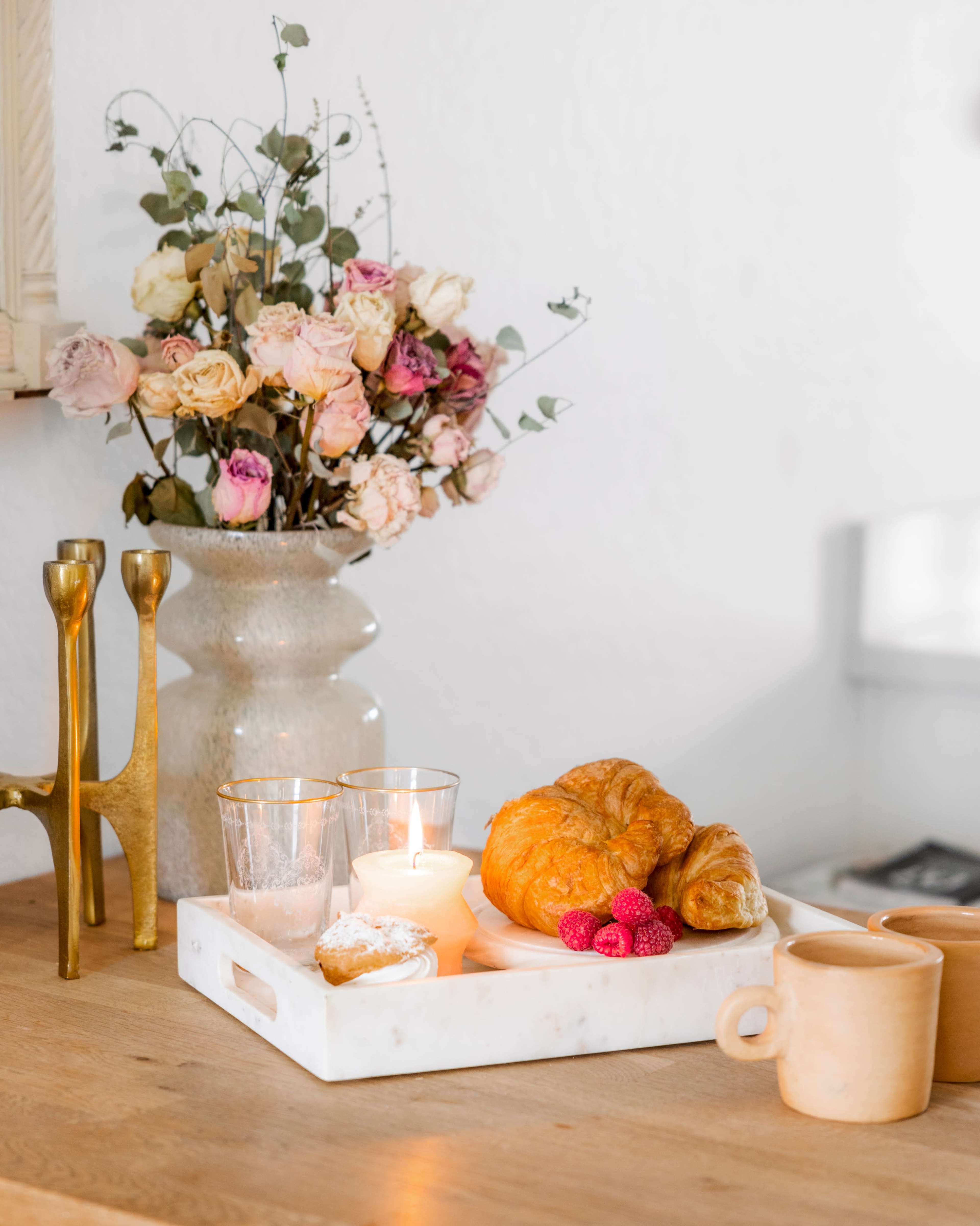 A wooden table holds a white tray with pastries, raspberries, candles, and a mug, alongside a vase of dried flowers and decorative candle holders.