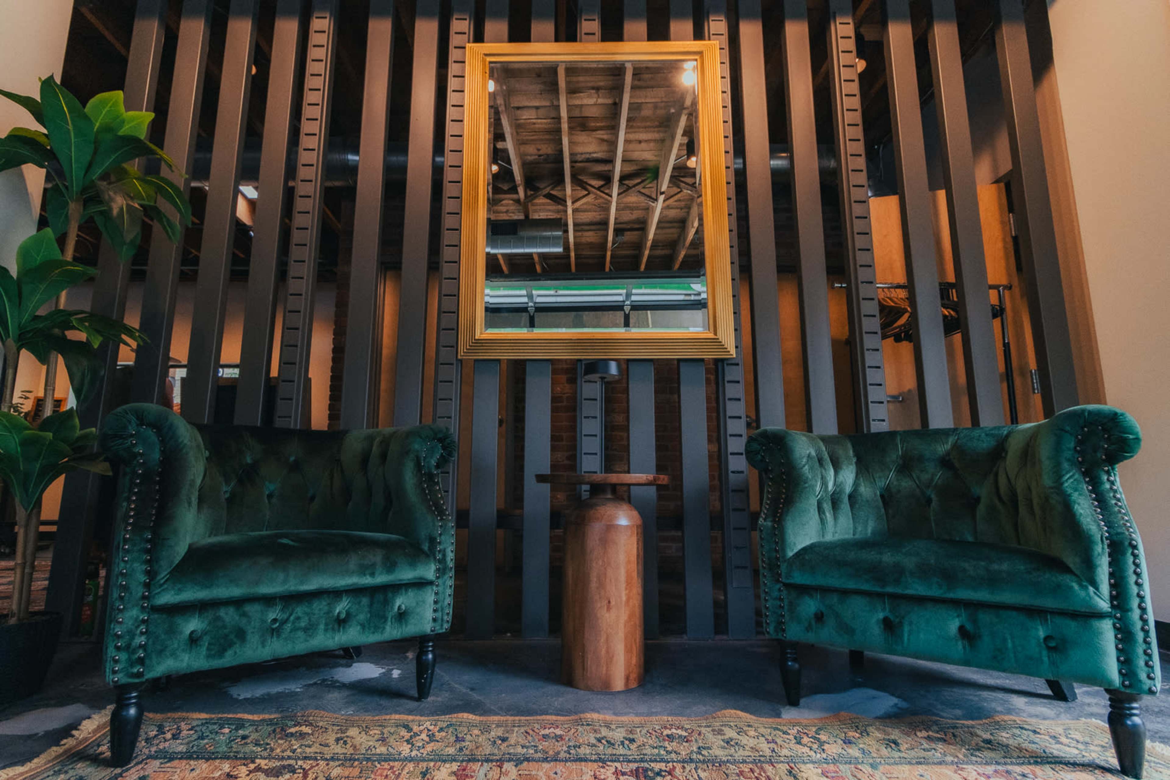 The image shows two green velvet chairs positioned opposite each other in front of a wooden wall, with a gold-framed mirror hung between them and a wooden side table in the center.