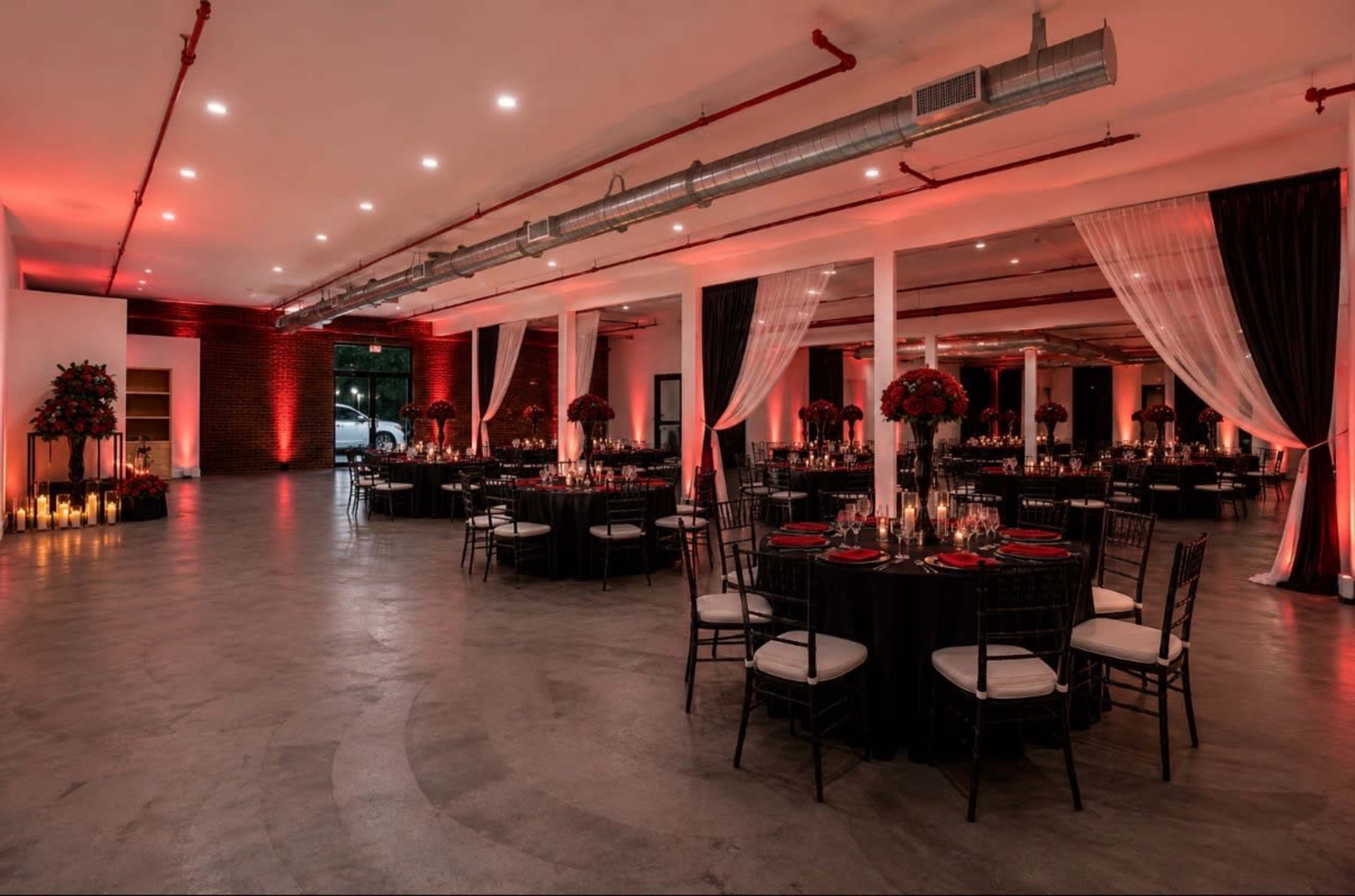 The image depicts a spacious, elegantly arranged event hall featuring round tables set with black tablecloths, red accents, and tall floral centerpieces, illuminated by soft lighting and candles.