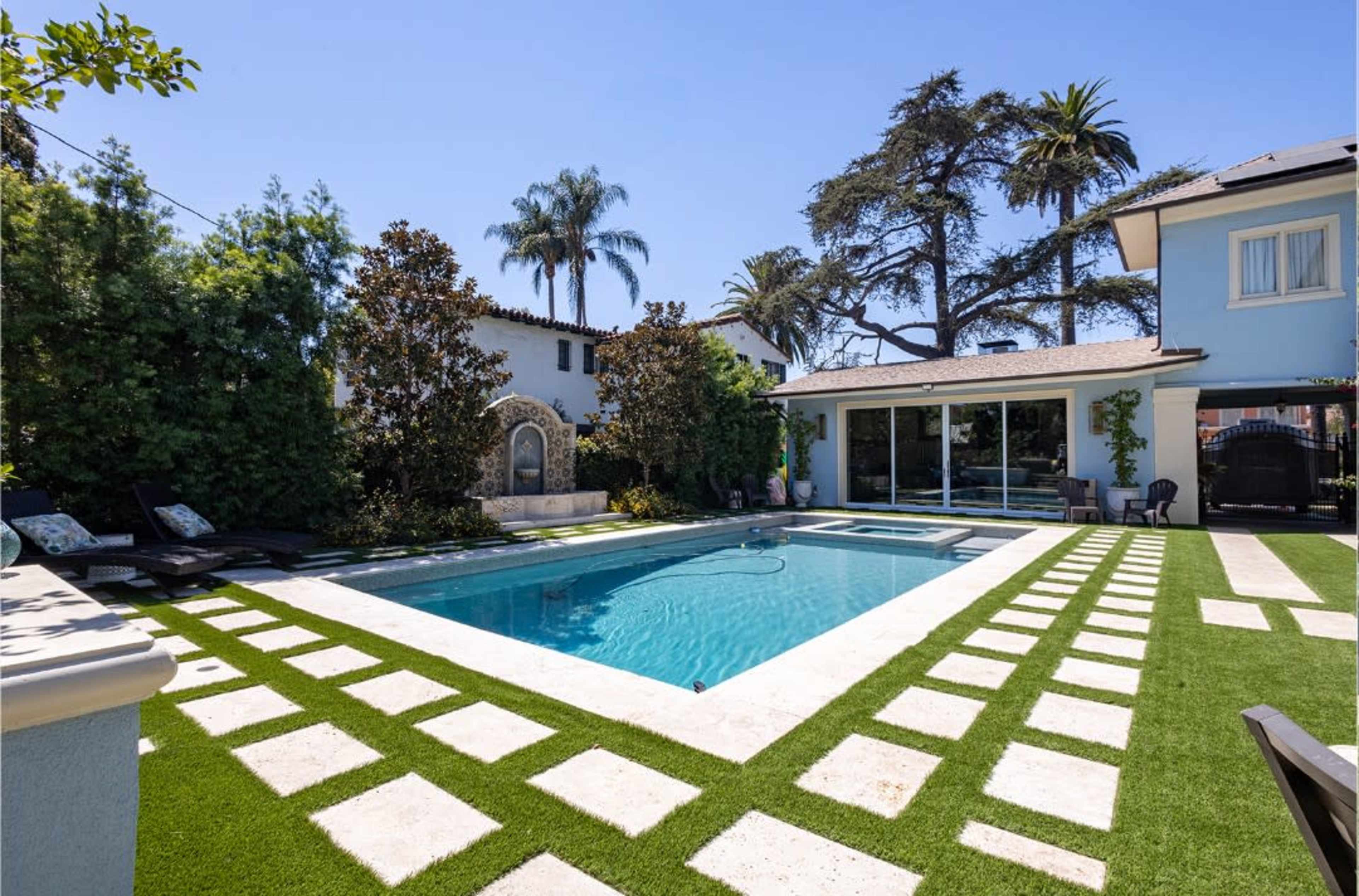 The image shows a backyard with a rectangular swimming pool surrounded by green grass and stone pathways, with palm trees and a house in the background.