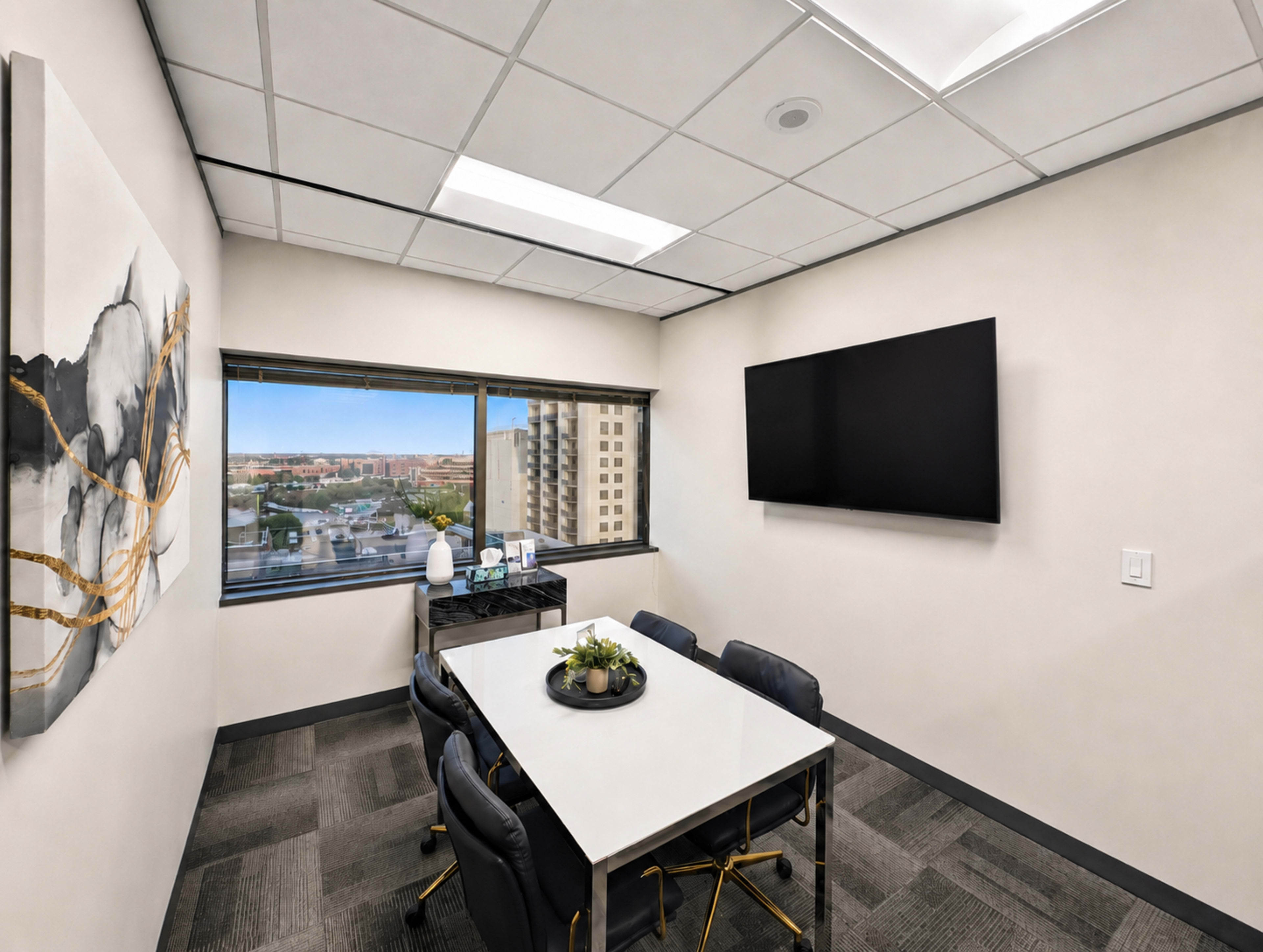 A small conference room features a white table with black chairs, a window overlooking a cityscape, and a mounted television on the wall.