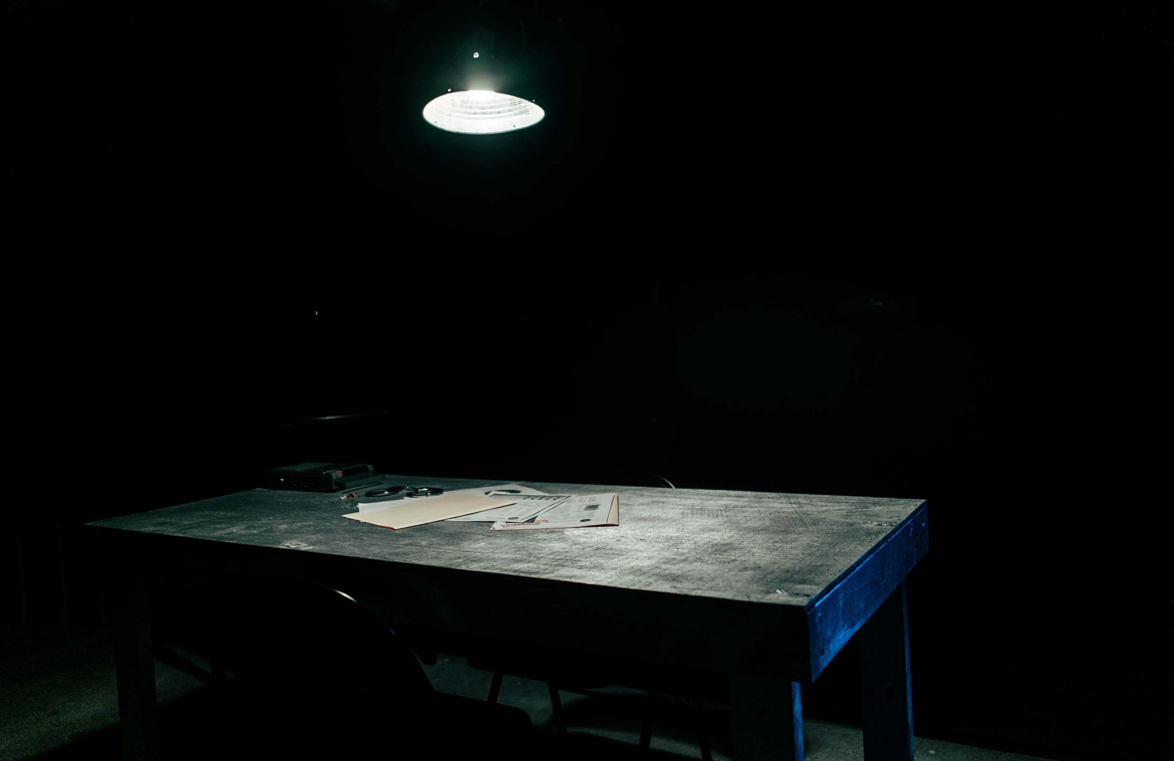 A wooden table with scattered papers and a phone is illuminated by a single overhead light in a darkened room.
